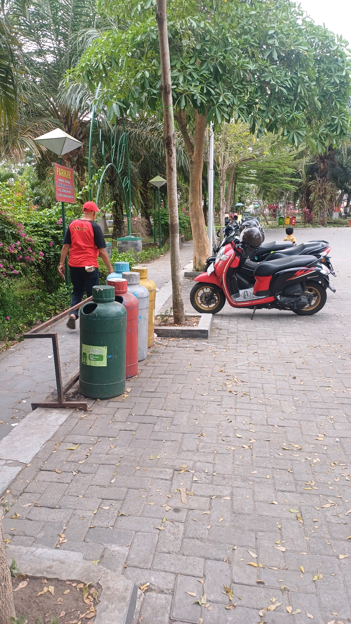 In Gergunung, Indonesia, a park scene unfolds with a person in a red shirt and cap walking away from the camera, heading towards a row of colorful trash bins. The bins, green, blue, red, and yellow, are lined up on a paved walkway bordered by trees and lush greenery. A red scooter with two helmets parked on it is visible further down the path, alongside other parked motorcycles. The ground is covered with scattered dry leaves, suggesting a mild season. A sign with "PARKIR" (parking) is posted in the background.