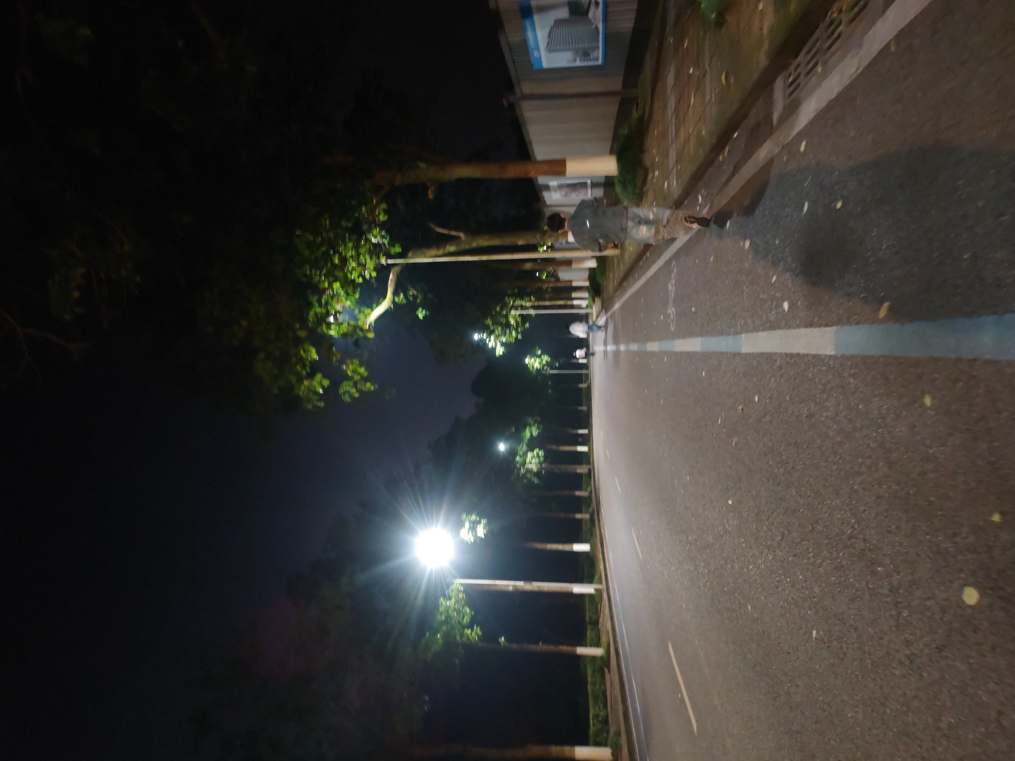 An eye-level photograph, apparently taken at night in Chongqing High-tech Zone, China, depicts a road receding into the darkness. The camera is positioned on the road or very close to it, looking toward the receding line of streetlights. The road, presumably asphalt, has a faint white line along its edge. A pedestrian is standing on the sidewalk on the right side of the road. The environment is characterized by a combination of urban infrastructure and natural elements. Trees line the road, their foliage obscuring the night sky. Streetlights cast pools of light along the road, with the closest one appearing as a bright, diffused light source. The background is dark, with the road appearing to narrow and disappear into the distance. A building is visible along the right side of the road.