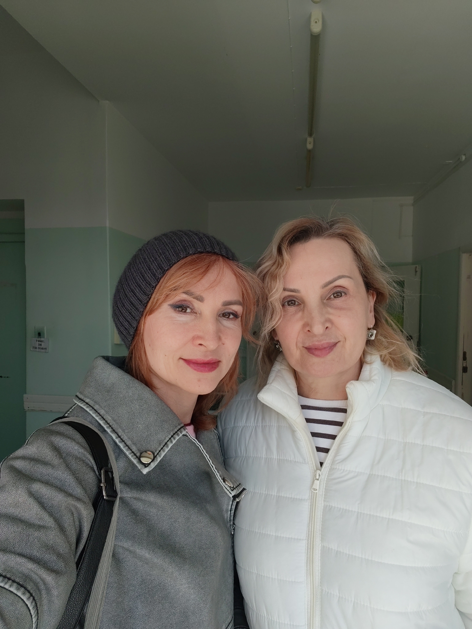 The image captures two women posing for a photo in an indoor hallway, likely in Chaikovsky, Russia. The woman on the left has reddish-brown hair and is wearing a dark grey knitted beanie and a grey faux leather jacket with silver snap buttons. A black shoulder bag strap is visible on her left shoulder. She has subtle eye makeup and pink lipstick. The woman on the right has wavy blonde hair and is dressed in a white puffer jacket over a black and white striped long-sleeved shirt. She wears distinctive silver square earrings. Both women are smiling gently and looking directly at the camera, suggesting a selfie.
The background shows a hallway with walls painted in two tones: a light mint green on the lower half and white on the upper half. The white ceiling features elongated fluorescent tube light fixtures. A door, also light-colored, is visible to the left. Text on this door appears mirrored, a common effect in selfies. When unmirrored, the bottom line clearly reads "БОЛЬШАЯ" (Bolshaya), meaning "Large" in Russian, while the top line starts with a "T" followed by other partially visible characters. The women's attire suggests cool weather. The overall setting, with its functional lighting and paint scheme, resembles a public or institutional building's corridor.