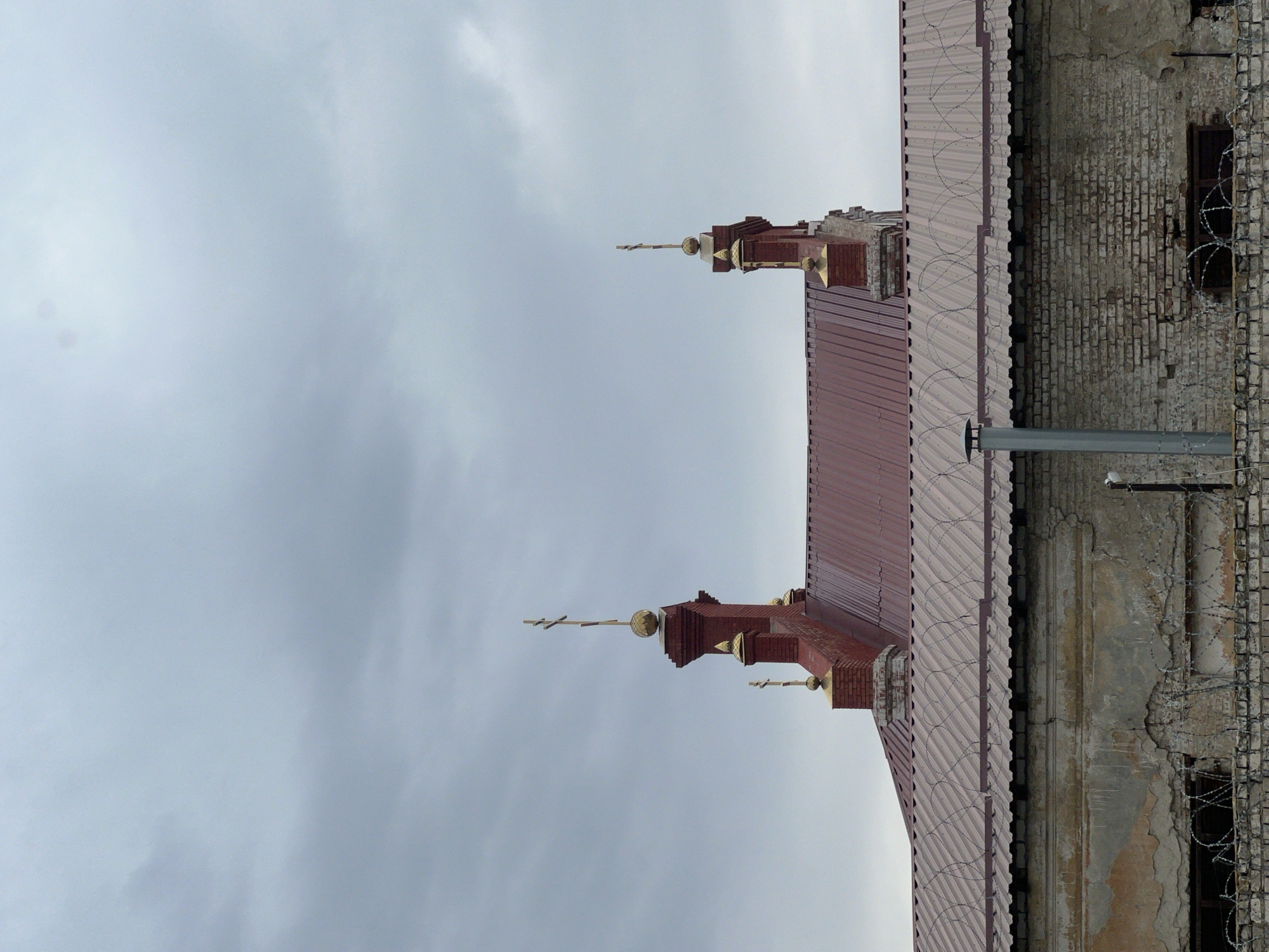 The image shows the roof and upper part of a brick building, likely a church or religious structure, against a cloudy sky. The building has a red corrugated metal roof with gold-colored crosses on top of brick towers. In the foreground, a lower section of a lighter-colored brick building is visible, with peeling plaster and barbed wire. A metal pipe or vent protrudes from the lower roof. The overall impression is one of a utilitarian structure combined with religious architecture, possibly in an urban or industrial setting.
The provided metadata indicates this is an image from Volgograd, Russia.