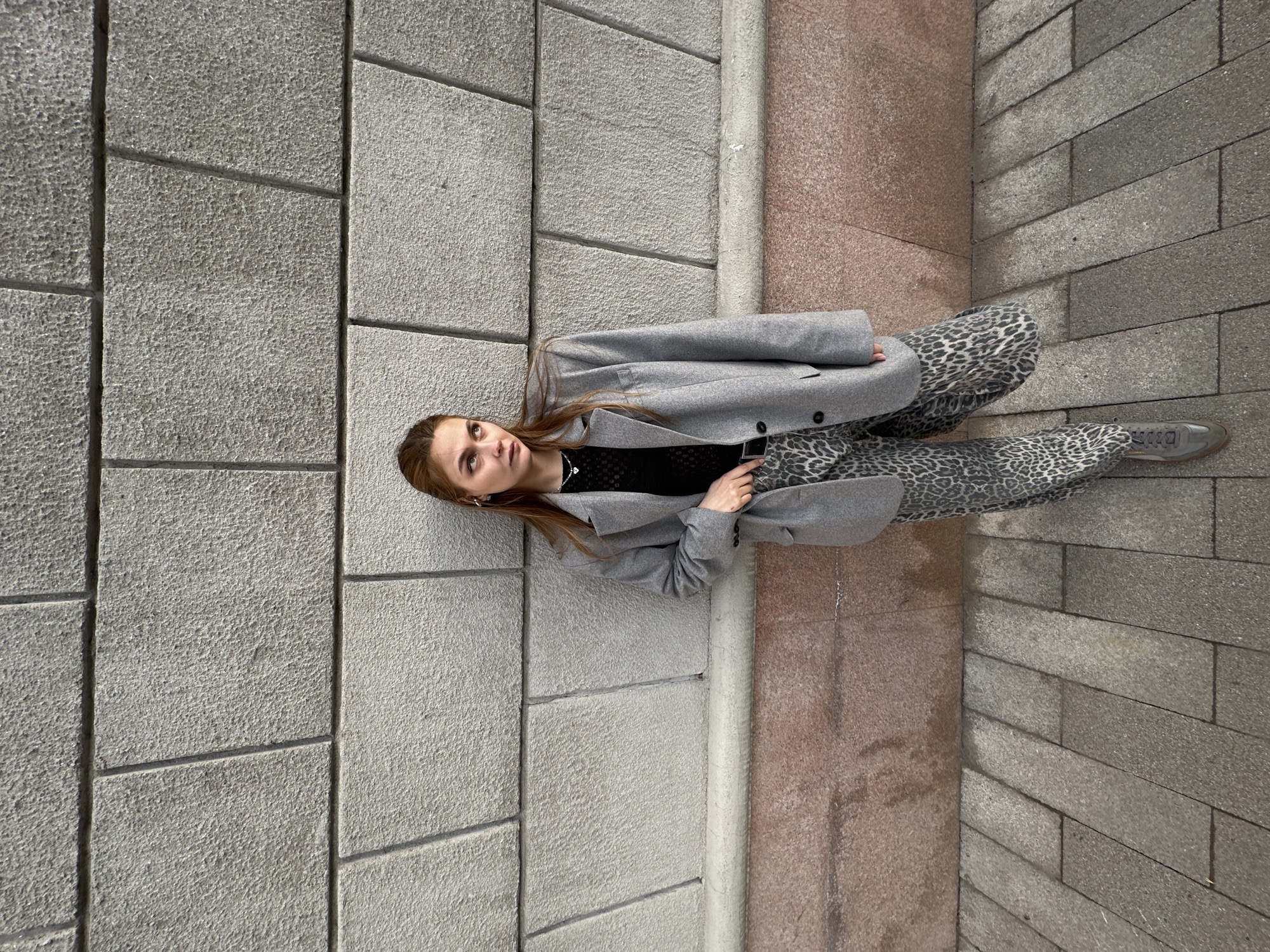 The image depicts a young woman posing outdoors. She is wearing a grey coat, a black lace top, and leopard-print pants, with grey shoes. She is lying on a paved surface consisting of large rectangular blocks and a strip of pink stone. She is looking upwards with a slight smile. The location is likely an urban setting with stone or concrete paving, possibly Krasnoyarsk, Russia. The image is taken from a top-down perspective.