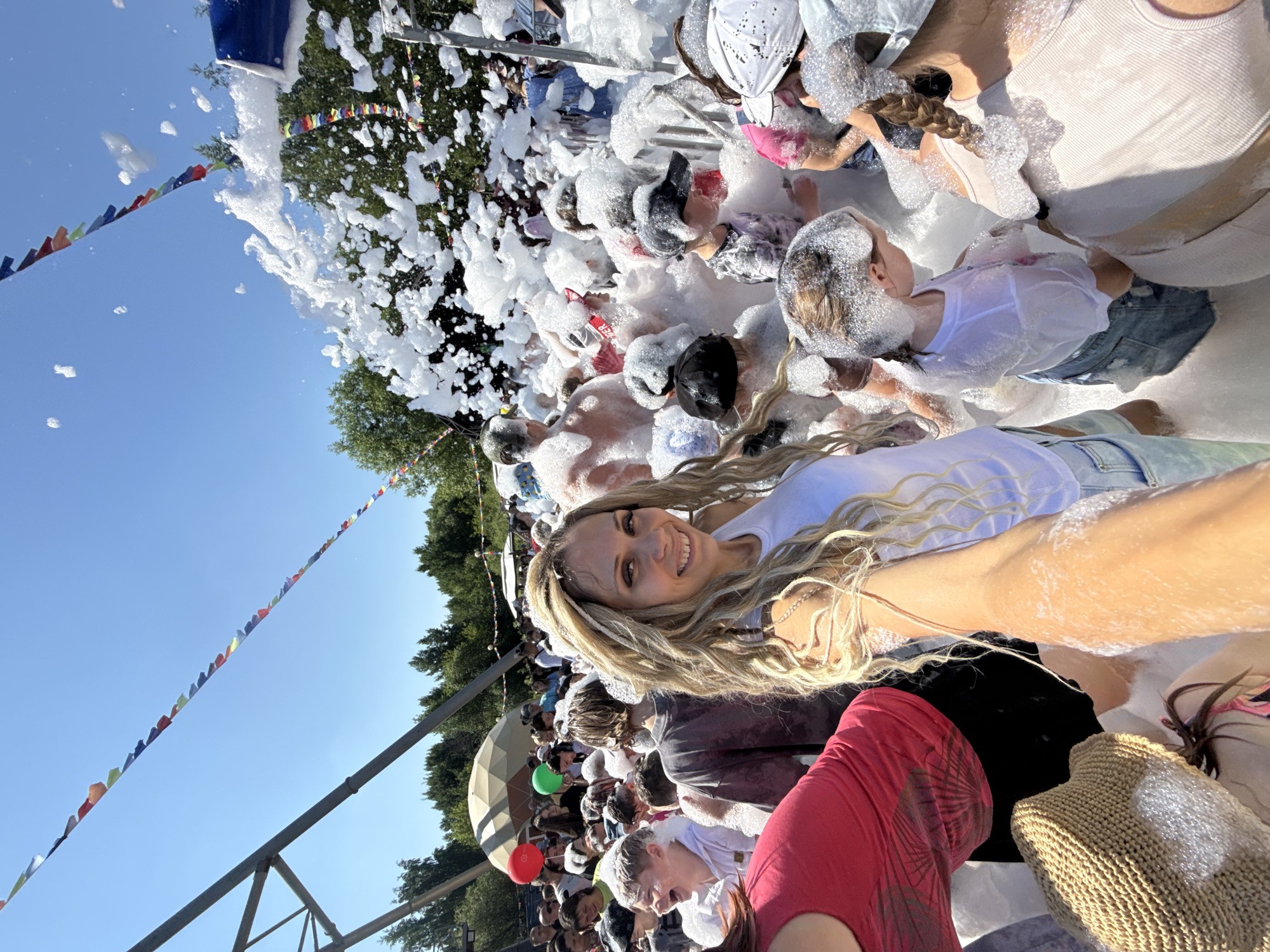 The image shows a sunny outdoor event, likely a foam party, in the city of Zavolzhye, Russia. In the foreground, a woman with long, blonde, braided hair smiles at the camera, her arm outstretched as if taking a selfie. She is wearing a white tank top and denim shorts. She is surrounded by a crowd of people, many of whom are children, all covered in white foam. The foam is being sprayed from a large blue machine visible in the upper right portion of the image. In the background, there are trees, a dome-shaped structure, and colorful triangular flags strung across the sky. Some individuals are holding red and green balloons. The overall atmosphere appears to be festive and playful.