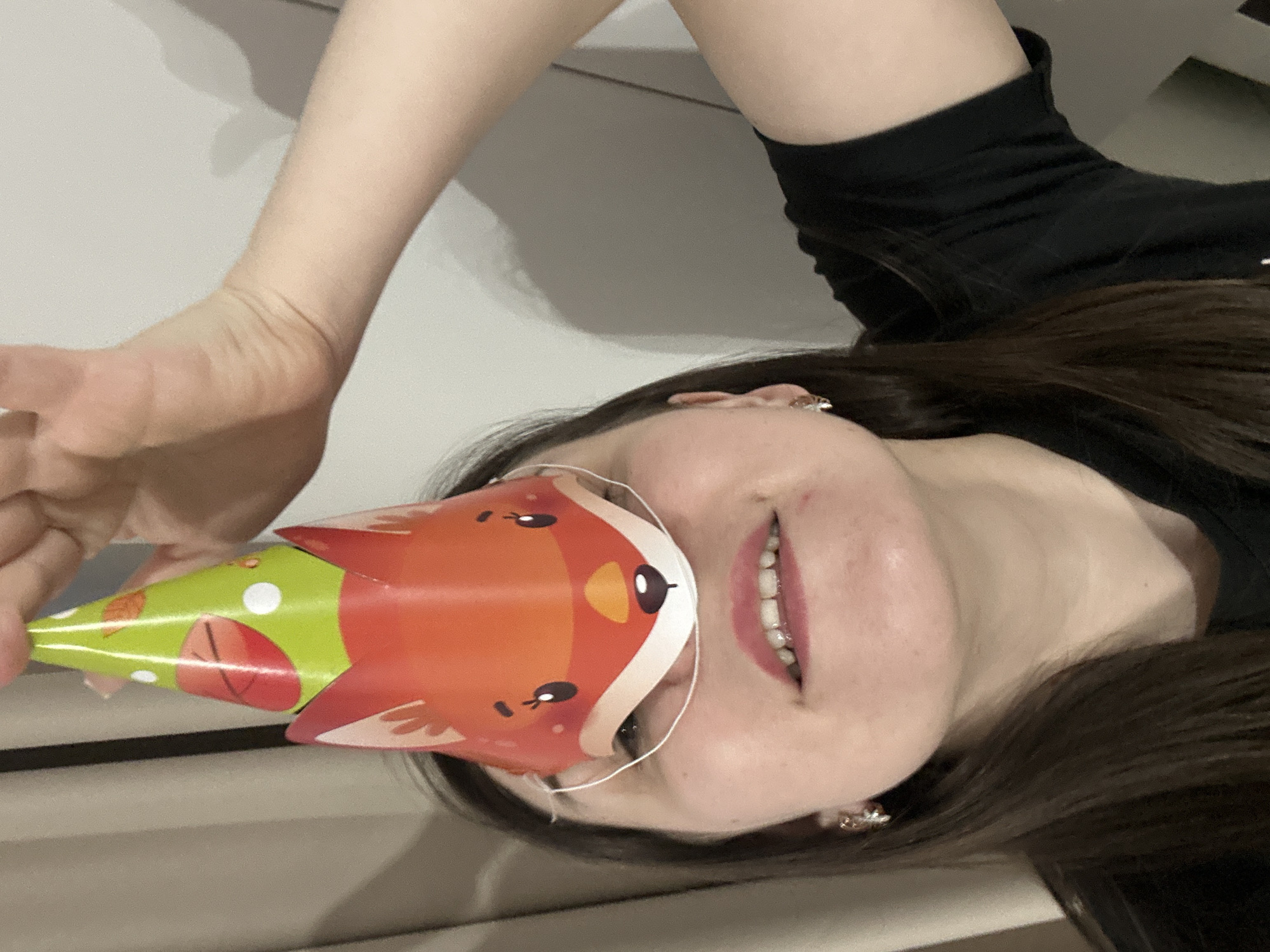 A person is holding a cone-shaped mask of a fox up to their face. The person has dark hair, light skin, and is wearing a black shirt. The mask is orange and white with a fox face printed on it. The background is a white wall. The location is not specified in the image and cannot be determined.