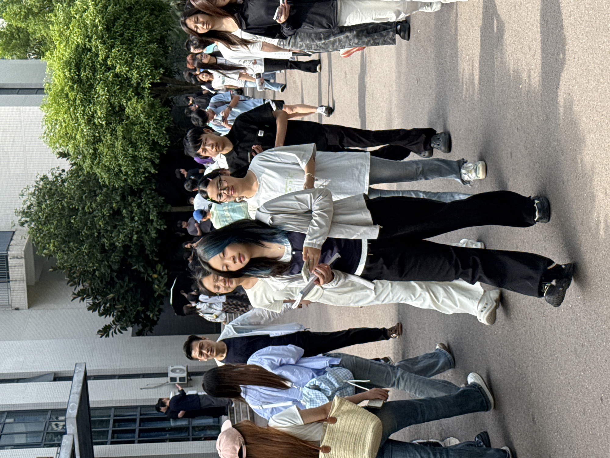 A long line of people, mostly young adults, stands on a paved surface alongside a building and a tree, possibly in Jiangjin, China. The people are dressed in varied casual clothing, including jeans, black pants, t-shirts, and blouses. Some are holding phones or papers. The building has windows and an air conditioning unit visible. The tree is full of green foliage. The people appear to be waiting in line.
