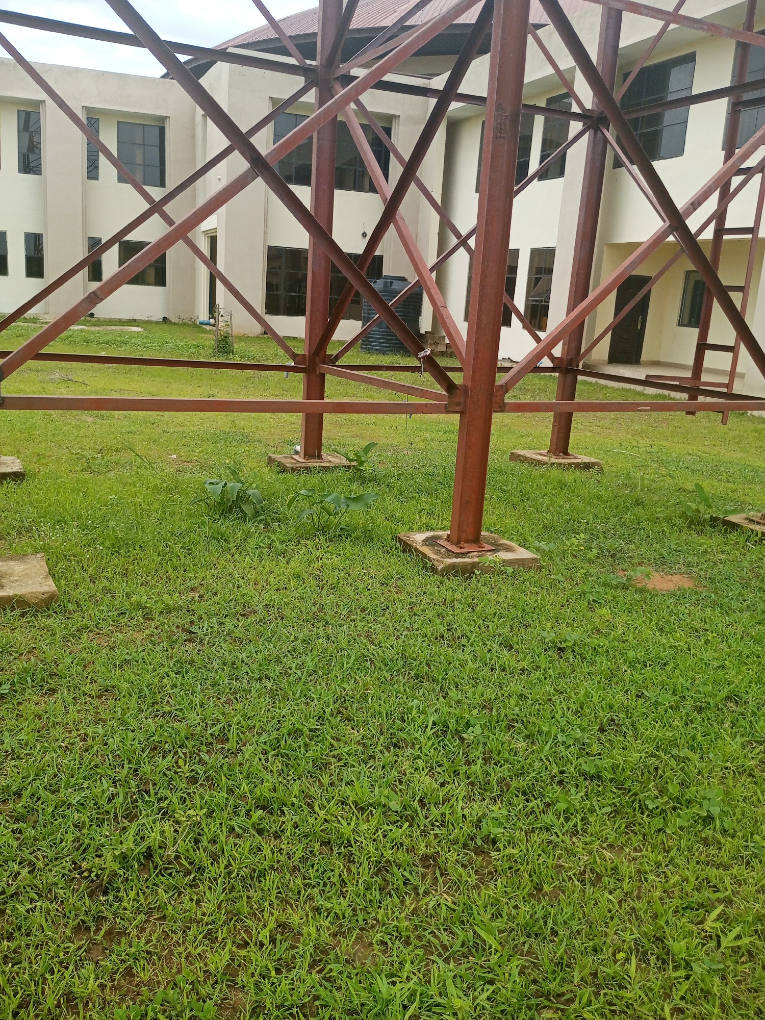 The media file depicts an outdoor scene in Jalingo, Nigeria, featuring a partially constructed metal framework in the foreground. This framework appears to be part of a larger structure, possibly a tower or a support system, with thick red metal beams forming an intricate lattice pattern. Three prominent vertical metal posts, also painted red, are anchored to concrete bases on the grassy ground.
Behind the metal structure, a modern, multi-story building is visible. The building has a light-colored facade and features numerous dark-tinted windows, suggesting it is a commercial or institutional building. A large black water tank is situated near the building, to the left of the metal framework. The ground in front of the building and around the metal structure is covered in lush green grass, with some patches of bare earth visible. Several small plants are growing in the grass. The scene appears to be set during daylight, with an overcast sky suggesting possibly cloudy weather. The overall impression is one of ongoing construction or an undeveloped area adjacent to a completed building. There are no people visible in the frame. No discernible text is present in the image.
