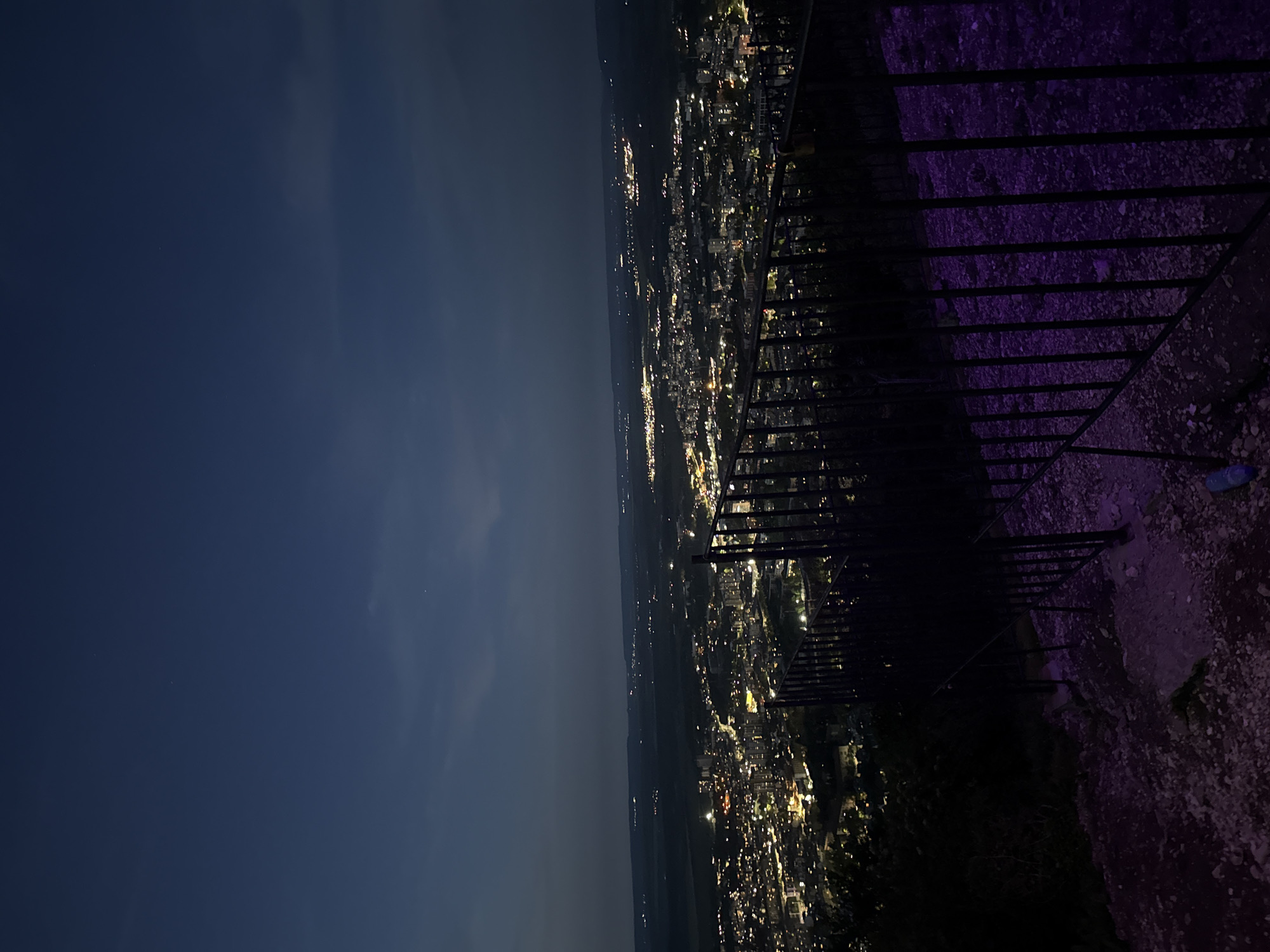 The image captures a nighttime view of Shumen, Bulgaria, from a high vantage point. The city sprawls across the landscape, illuminated by numerous lights that delineate streets and buildings. A dark sky covers the city. A fence runs along the right edge of the image. The platform on which the fence is built is rocky and uneven. The lighting and perspective suggest the photograph was taken from a high overlook in Shumen.