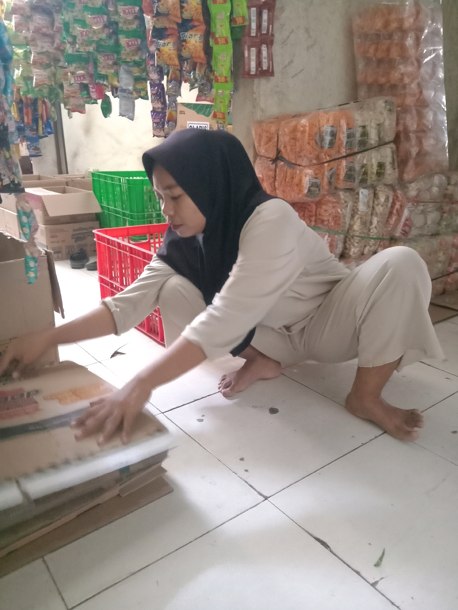In Sugihwaras, Indonesia, a woman wearing a black hijab and light-colored clothing is crouched on a tiled floor, actively sorting through a stack of flat cardboard items. Her left hand is positioned on top of the stack, suggesting she is organizing or examining them. In the background, various items are visible, including stacked boxes, green and red plastic crates, and rows of packaged snacks hanging from a rack, indicating the scene likely takes place in a small shop or market.