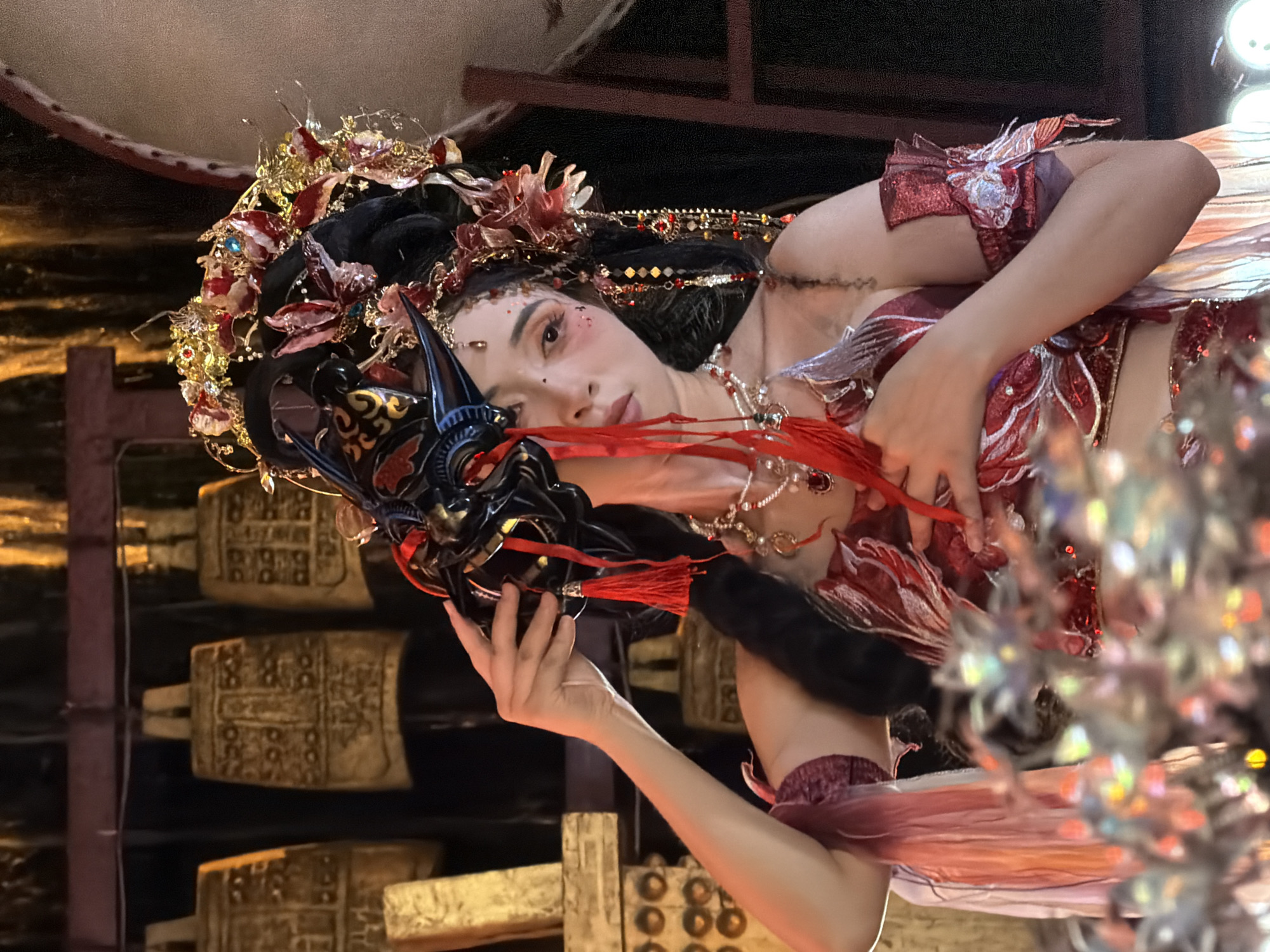 A woman in elaborate traditional Chinese costume, including a mask and headdress, is holding the mask to her face. She is wearing a red and gold outfit adorned with feathers and beads. Her makeup is dramatic, with red blush and glitter. Behind her, several large, ornate bells are hanging from a wooden structure. The scene appears to be set indoors, with warm, golden lighting. The overall impression is one of theatricality and cultural performance. The location is not specified in the image or the provided crops.
