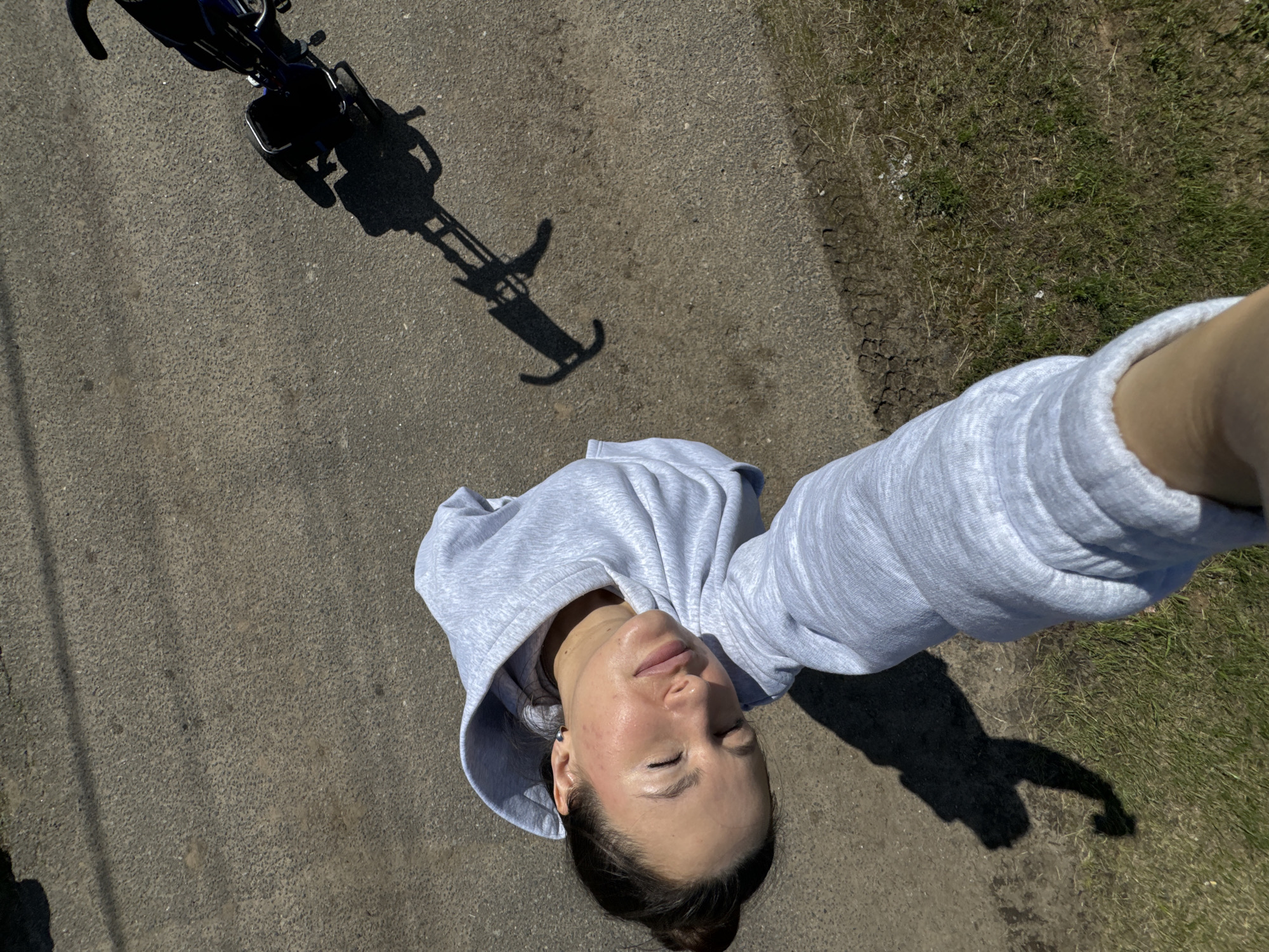 A woman in a gray hoodie lies on a paved surface near a grassy area, taking a selfie with her arm extended. A bicycle and its shadow are visible in the upper left corner of the frame. The scene appears to be outdoors on a sunny day. Location cannot be determined.