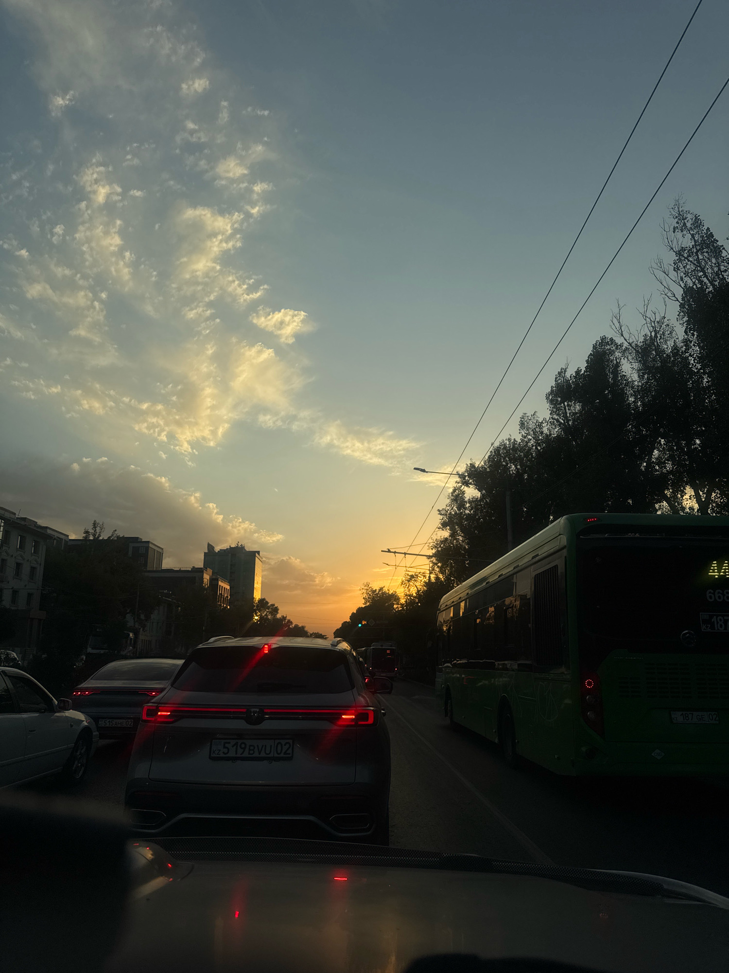 A street-level view captures a traffic jam during sunset on a city street in Almaty, Kazakhstan. The sky displays a gradient of warm oranges and yellows from the setting sun, illuminating scattered clouds. In the foreground, the rear of a silver SUV is visible, its brake lights illuminated. To the right, a green bus with the number "668" displayed prominently on its front moves in the same direction. The bus's license plate reads "187 GE 02". A traffic light is visible in the distance, glowing green. Tall trees line the right side of the road, their dark silhouettes contrasting with the bright sky. Buildings, including apartment complexes and taller structures, are visible on the left side of the street. Overhead, power lines stretch across the sky. The overall impression is a typical urban commute at dusk, with the natural beauty of the sunset creating a striking backdrop.