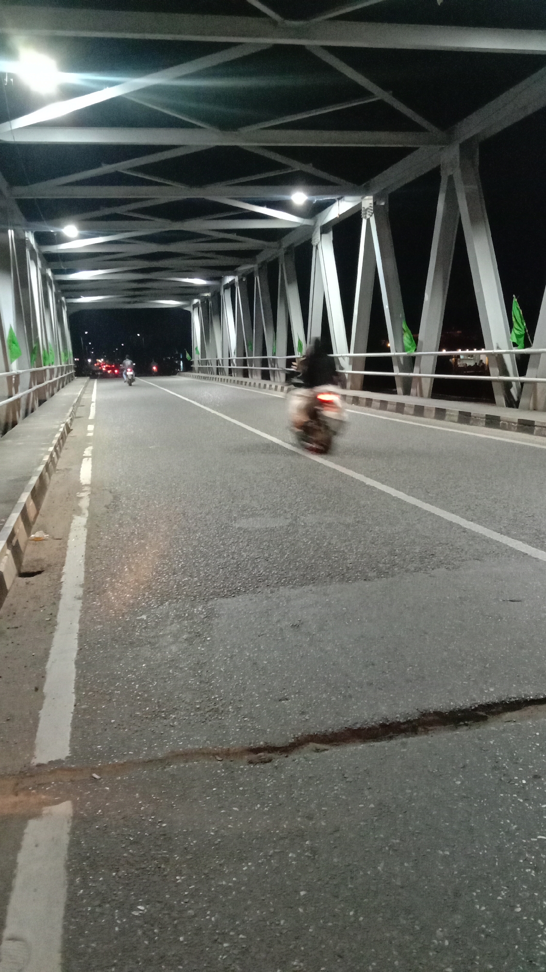 The image shows a bridge at night in Lhokseumawe, Indonesia. The bridge has a steel frame structure with overhead lighting. A white line marks the center of the road. On the bridge are two motorbikes, one moving away in the right lane, and another smaller one further away in the left lane. Green flags are attached to the side railings.