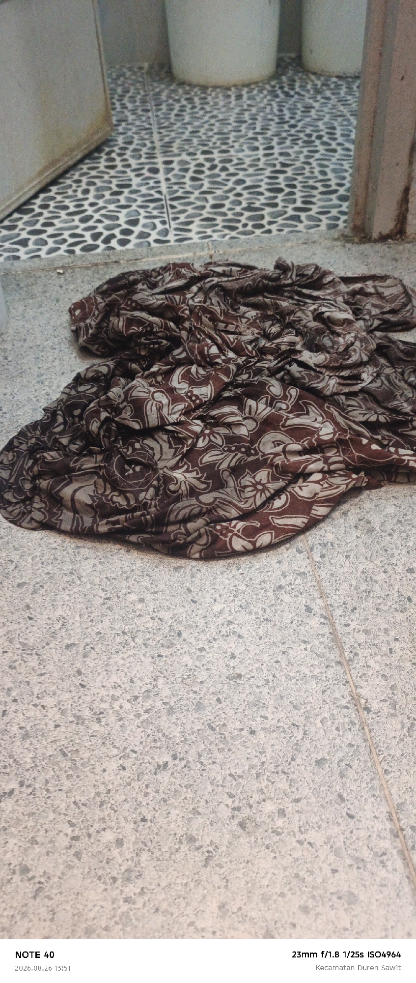 A crumpled dark brown and tan patterned cloth lies on a speckled gray floor. The cloth has a repeating floral or paisley-like design. Behind the cloth, there is a tiled area with a black and white pebble-like pattern, and further back, two large white cylindrical objects are visible, possibly buckets or planters. The overall environment appears to be outdoors or in a covered outdoor space, given the flooring and the presence of what looks like a wall or large structure to the right. The bottom of the image shows text overlays indicating "NOTE 40", a date "2026.08.26 13:51", and camera settings "23mm f/1.8 1/25s ISO4964", along with the location "Kecamatan Duren Sawit", which is a subdistrict in East Jakarta, Indonesia.