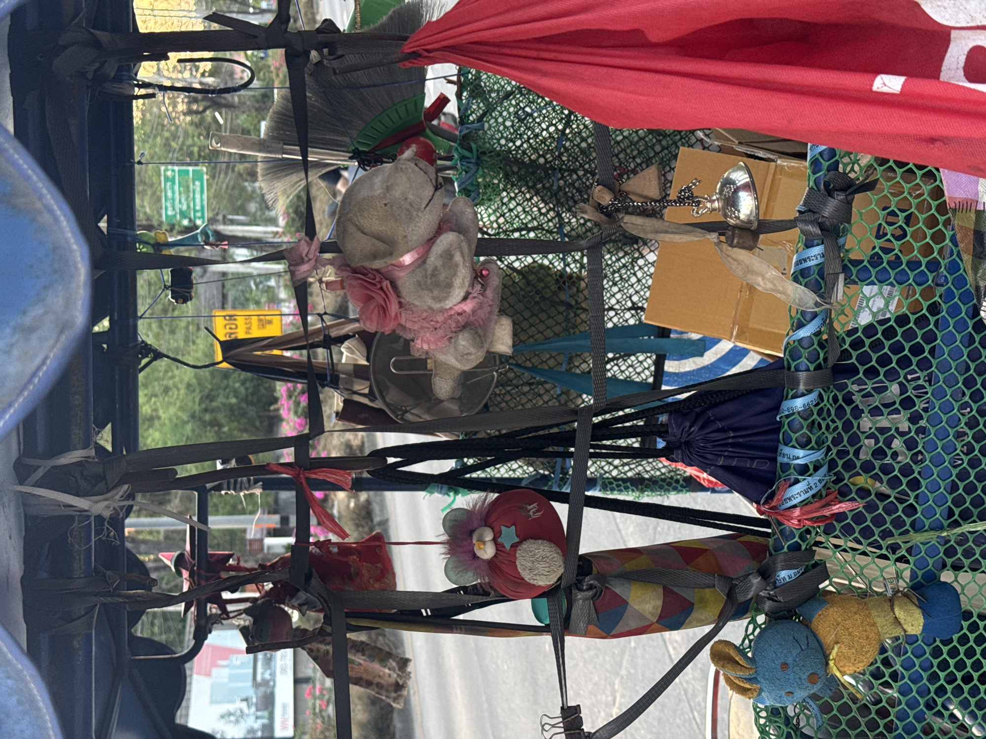 The image depicts a cluttered rack of miscellaneous items, possibly attached to a vehicle in บ้านกาญจน์กนก 23, Thailand. The items are secured with straps and include:
* Stuffed animals (a bear with a pink ribbon and flower, a red heart-shaped plush with embellishments, and a blue/yellow plush)
* Brooms
* A red cloth or fabric
* A silver chalice or ornament
* A cardboard box
* A green and blue net
* Drawstring bags
The rack is made of a dark metal frame. The background shows a street with buildings and vegetation, suggesting an urban environment. The overall impression is one of disorganized storage or transportation of goods.