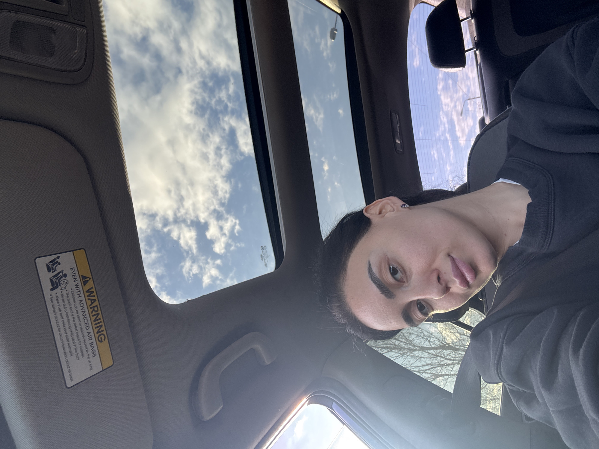 The image is taken from inside a car, presumably a selfie. The interior includes a sunroof with a view of a partly cloudy sky. A woman is visible in the passenger seat, wearing a black sweatshirt. The car's sun visor is visible, featuring a warning label about airbags. The location is unknown and there is no information to suggest it is Tbilisi, Georgia.