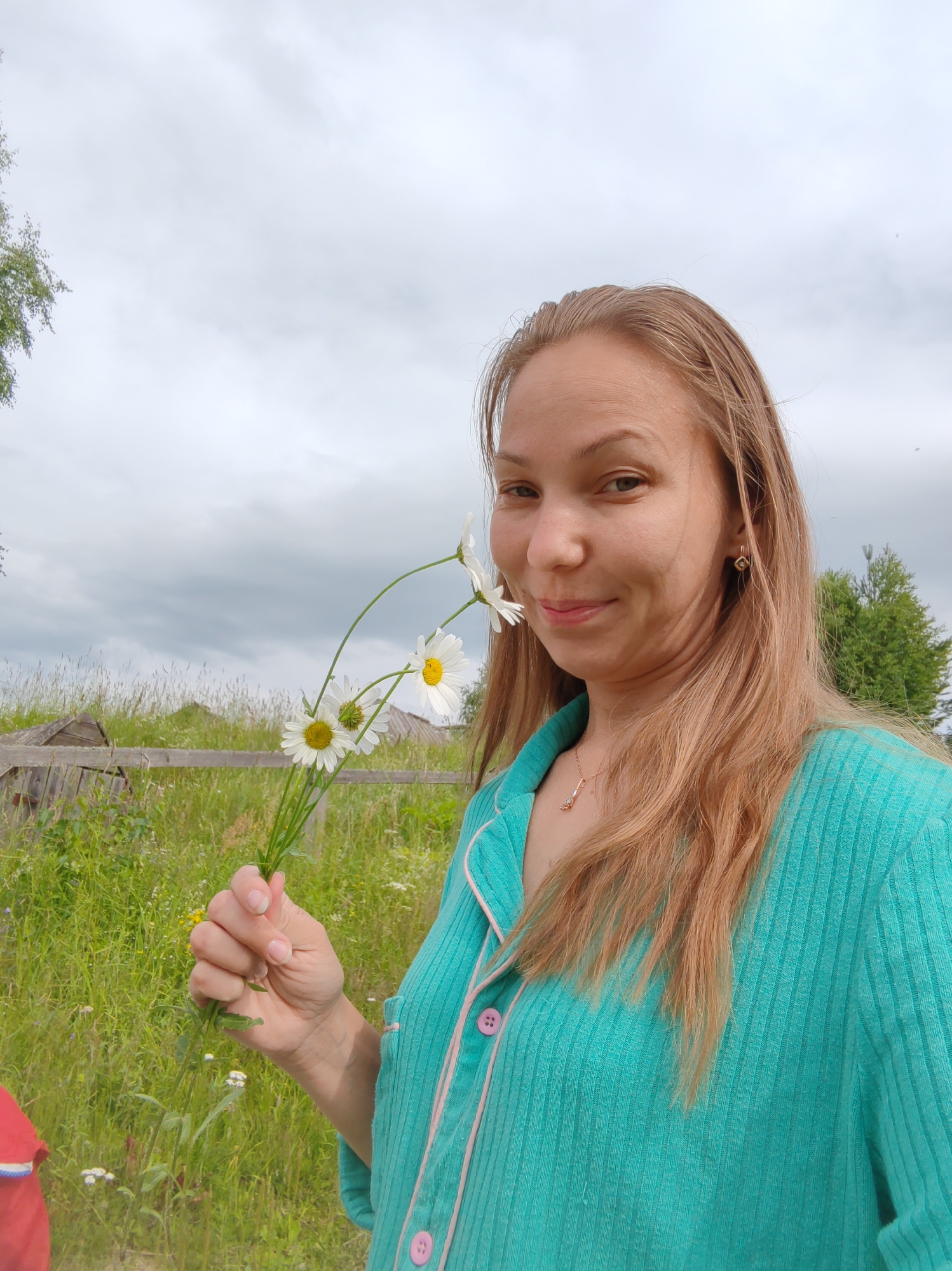 A young woman with long, blonde hair is taking a selfie outdoors. She is holding a small bouquet of daisies. She is wearing a teal colored pajama-like top with pink trim and buttons. A small, gold pendant is visible around her neck. In the background, there is a wooden fence and tall grass, suggesting a rural or semi-rural setting. The sky is overcast and grey. The location is the city of Chukhlem, Russia.