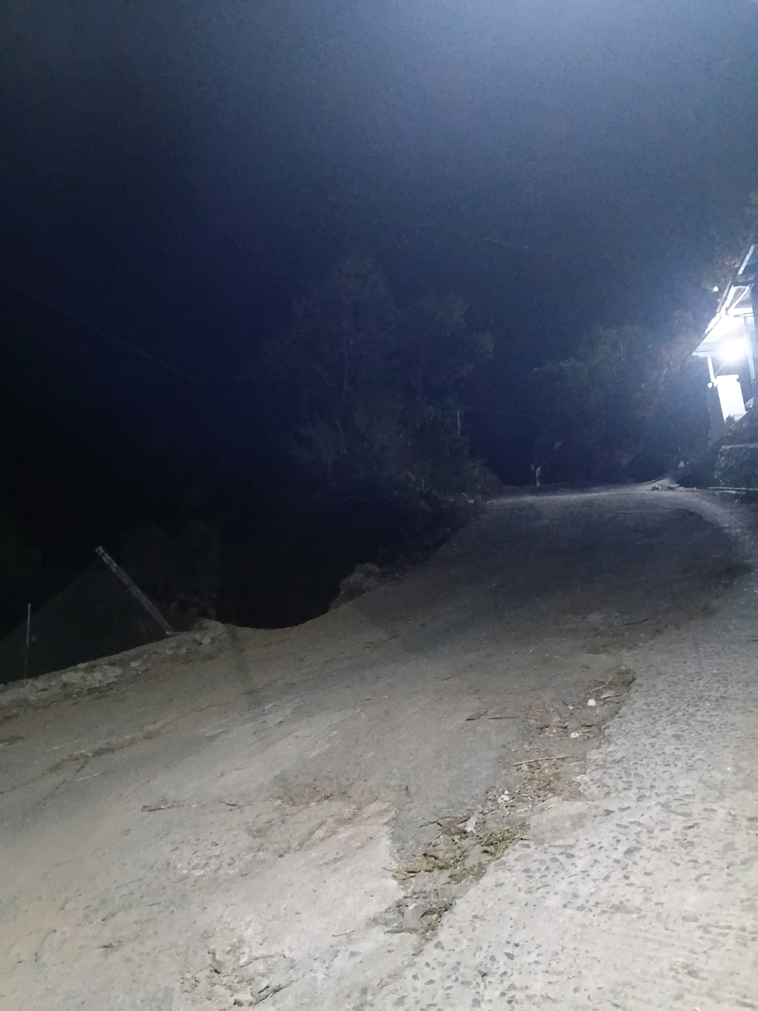 The image depicts a dimly lit, unpaved road at night. The road surface is uneven and appears to be made of compacted dirt and gravel, with some debris scattered across it. To the right of the road, there is a structure with a bright light emanating from it, illuminating a section of the road and a wall made of stone. To the left of the road, the terrain slopes upwards and is covered in vegetation and trees, with a dark, shadowy expanse beyond. A single, indistinct human figure can be seen standing further up the road, illuminated by the light from the structure. The overall impression is of a rural or semi-rural setting in the city of Sinduaji, Indonesia, at night.