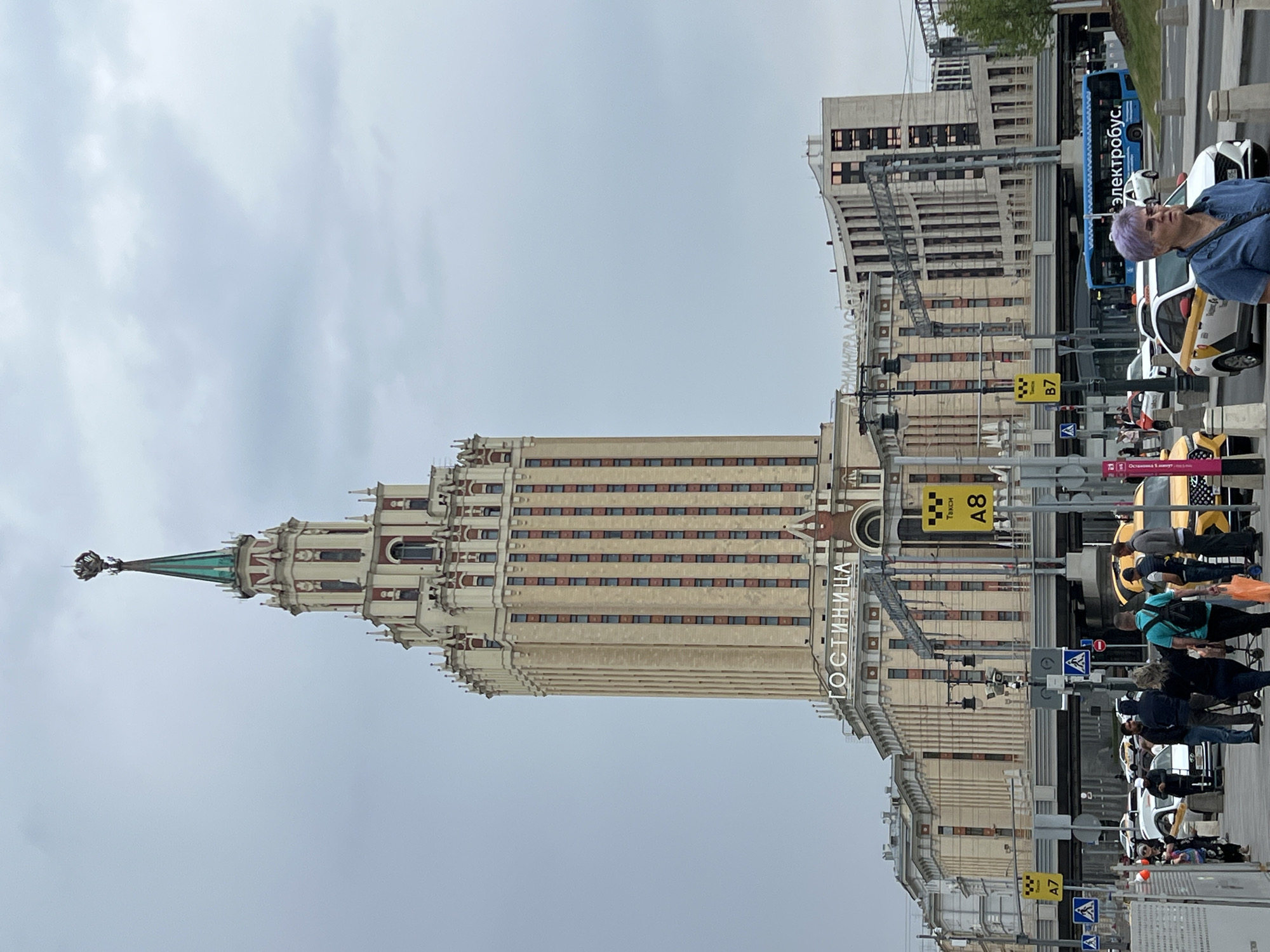 The image shows a view of the Hotel Ukraina (now Radisson Collection Hotel, Moscow) in Moscow, Russia, under a cloudy sky. The imposing Stalinist skyscraper, topped with a green spire and star, dominates the frame. Signs on the building read "ГОСТИНИЦА" (Hotel). In the foreground, a street scene unfolds with several yellow taxis, an electric bus labeled "электробус" (electric bus), and pedestrians. Some pedestrians are walking, while others appear to be waiting or interacting. The environment includes streetlights, traffic signs, and a pedestrian overpass. The overall impression is a bustling urban environment with iconic architecture.