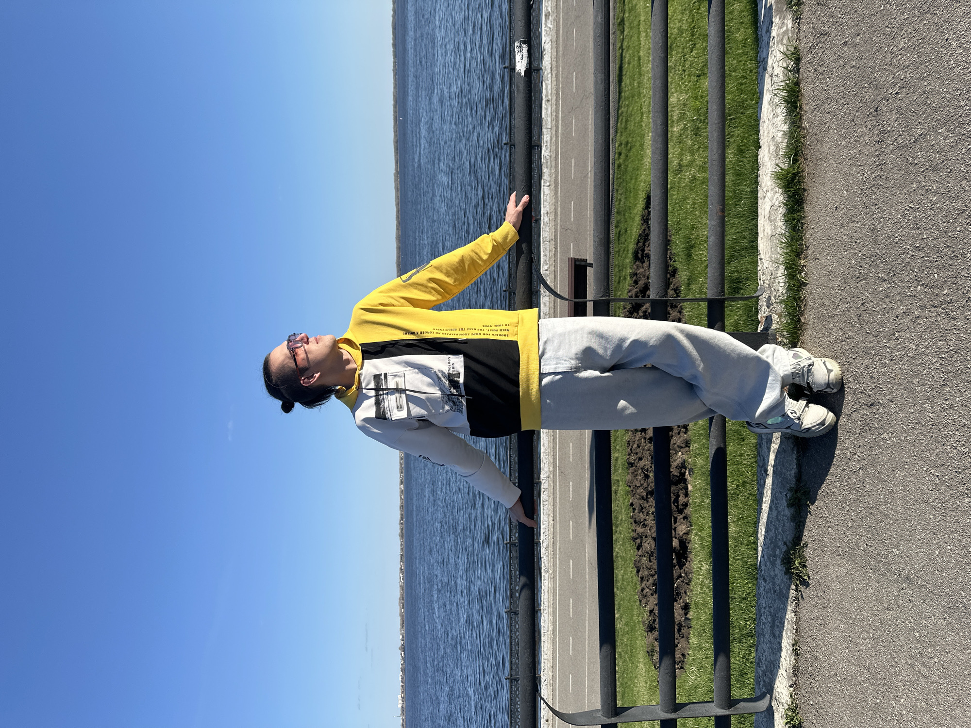 A person, likely male, is captured standing outdoors on a bright, sunny day in Saratov, Russia. They are wearing a distinctive yellow, black, and white hoodie, light gray pants, and white sneakers. Sunglasses rest on their face, and their hair is pulled back. The individual is looking upwards towards the clear, vibrant blue sky, with their left arm extended and resting lightly on a black metal railing.
The scene is set alongside a large body of deep blue water, likely a river or lake, which dominates the left side of the frame. Running parallel to the water is a paved road or path, visible beyond the black metal railing that separates it from the viewer's immediate space. The path features white dashed lines, indicating a road or lane. To the right of the railing is a strip of lush green grass, transitioning into an asphalt pathway where the person's feet are positioned. The strong sunlight casts clear shadows, suggesting it is midday.
Notable text fragments are visible on the person's hoodie: "LOOKING FOR HOPE FROM DESPAIR NO LONGER A BOLLOI MUCH WHA YOU HAVE THE ABILITYNECH TO COME MONE".