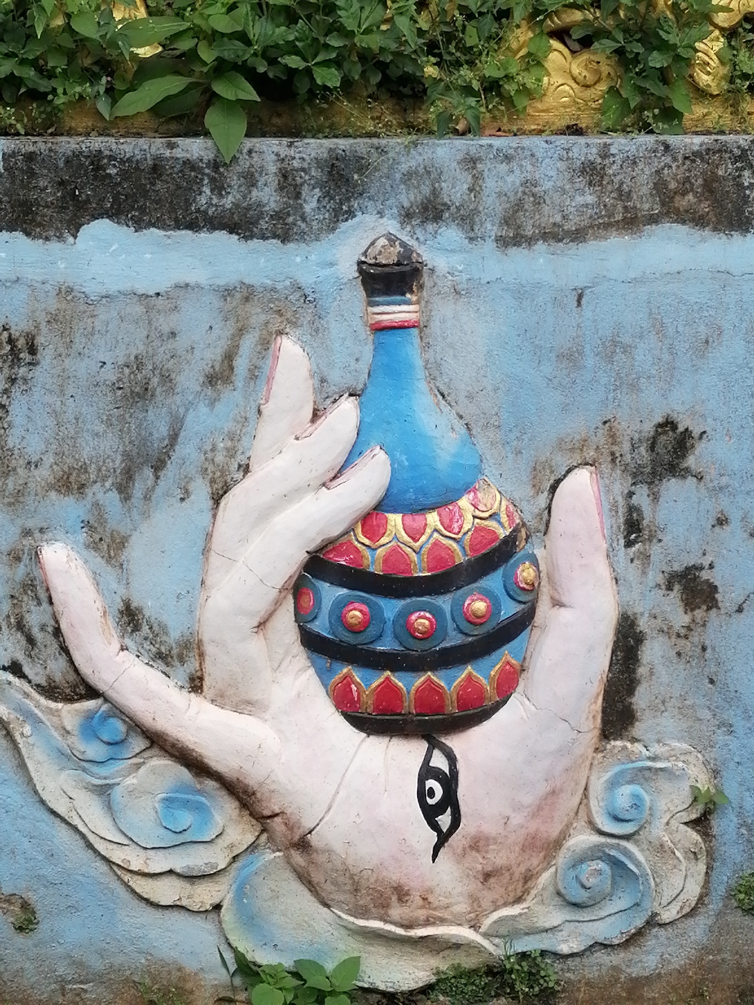 The image shows a close-up, low-angle shot of a relief sculpture on a weathered, light blue, concrete wall, likely located in Da Lat, Vietnam. The sculpture depicts an oversized, stylized, light-skinned hand with pink fingernails, cradling a blue, bulbous vase. The vase is decorated with a band of red and gold floral patterns, a black band with gold circles, and a blue band with red circular elements. Above the vase, the sculpture has a conical, dark top with red and blue banding. An eye symbol, in black and white, is painted on the palm of the hand. The hand rests on stylized, light blue and white clouds. Green foliage is visible at the top of the image, and some vegetation grows at the bottom. The concrete wall shows signs of age and discoloration, with dark staining and cracks. The overall impression is of a religious or decorative element in an outdoor setting, possibly on a temple or public monument.