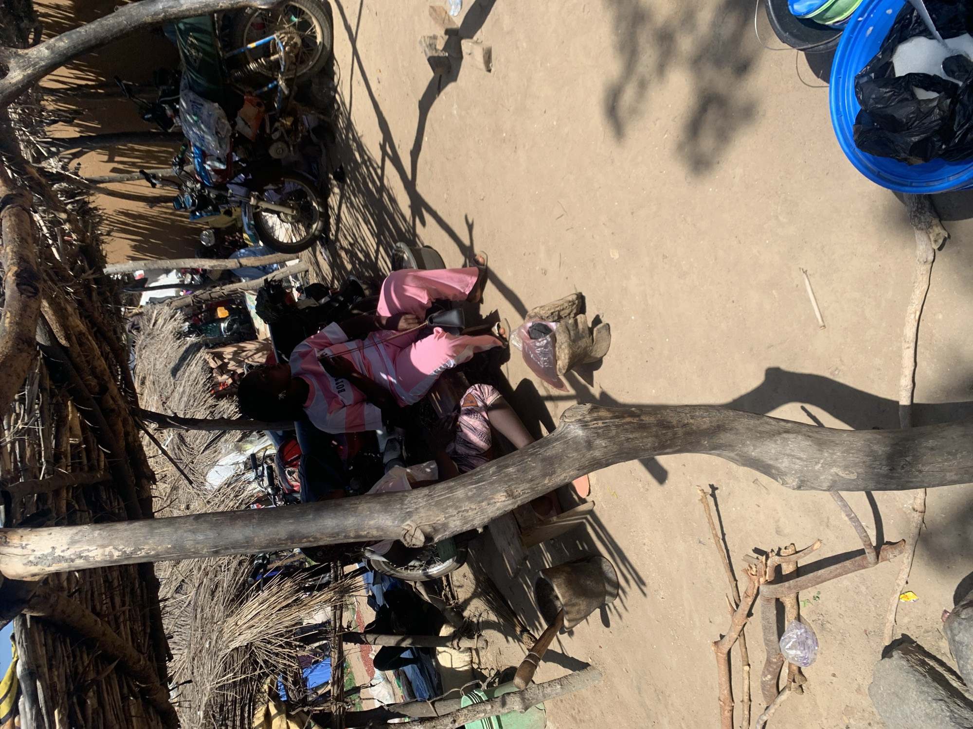 This media file shows an outdoor scene in Dull A, Nigeria. The image is oriented vertically. In the foreground, a large, weathered tree branch stretches horizontally across the frame, casting a shadow. To the left, a rustic structure made of branches and dried vegetation suggests a simple dwelling or market stall. Inside this structure, several motorbikes are visible, some with items piled on them.
Further into the scene, two individuals are seated on the ground. One person, wearing a pink shirt and dark pants, is leaning back. The other individual, also seated, wears a pink and white striped garment. They appear to be resting or engaged in quiet activity. Scattered around them are various objects, including plastic bags, what looks like a basket, and wooden items.
In the background, the ground is dusty and uneven. The bright sunlight indicates it is daytime, and the strong shadows suggest a sunny weather condition. To the right of the frame, a blue plastic bucket contains black plastic bags, and there are other buckets or containers nearby. The overall impression is of a rural or semi-urban setting in Nigeria, with a focus on everyday life and the presence of motorbikes, which are common modes of transportation. The scene appears peaceful, with the individuals resting in the shade. No visible text is discernable in the image.