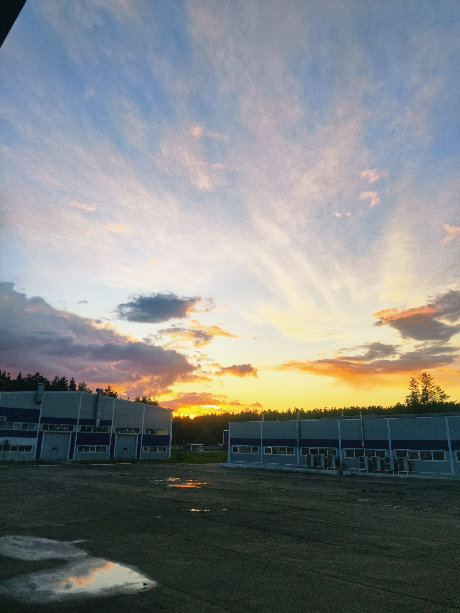 The image captures a twilight scene in Noviy, Russia, featuring two large, industrial-style buildings with gray facades and blue accent trim along the top and below the windows. The buildings are situated on a flat, paved surface with puddles reflecting the colorful sky. In the background, a dense line of trees is visible, silhouetted against the sunset or sunrise sky. The sky is a mix of blues, purples, oranges, and yellows, suggesting either dawn or dusk. No people or animals are visible in the image. Air conditioning units are seen along the base of the building on the right.