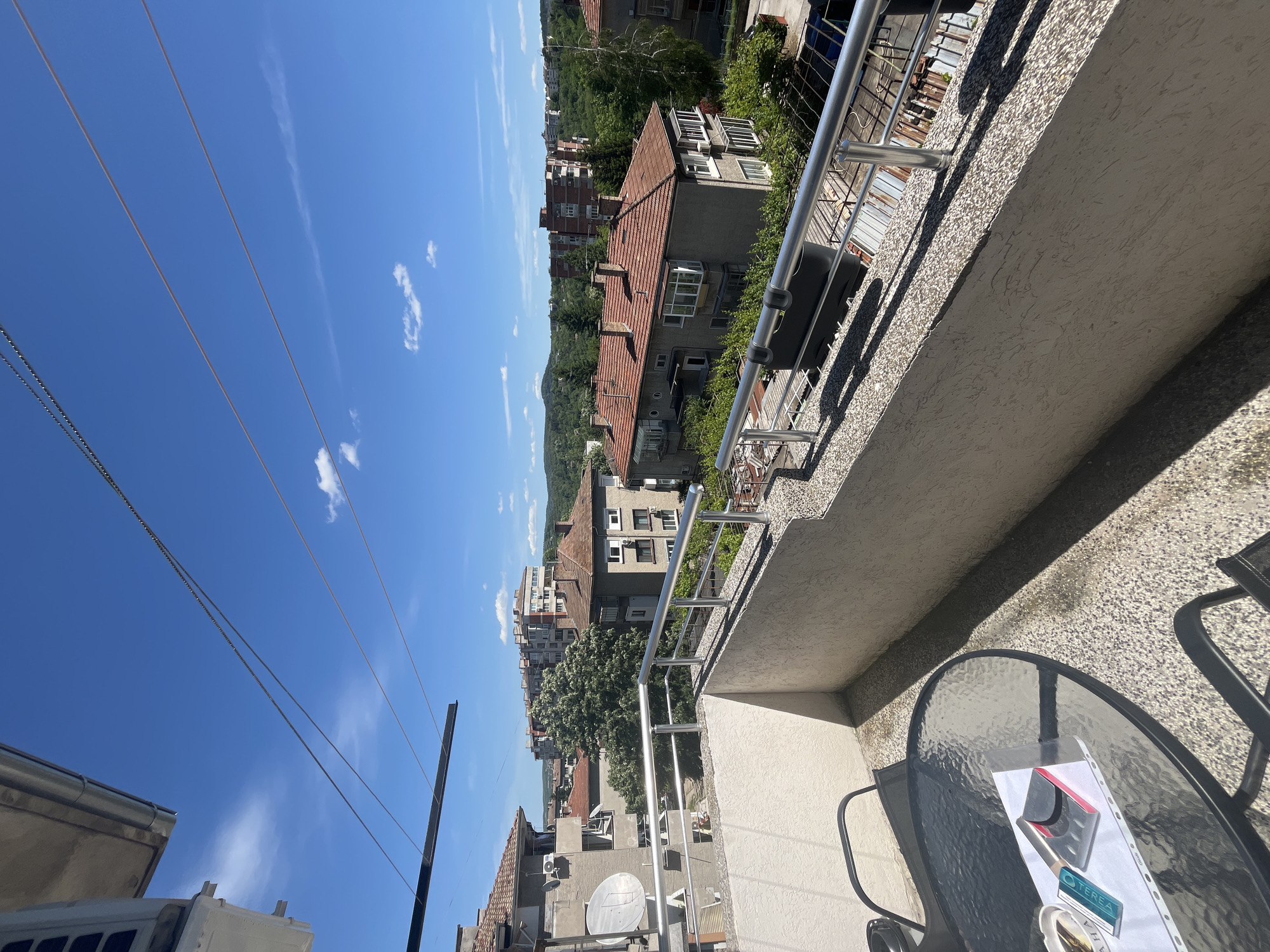 View from a balcony in Veliko Tarnovo, Bulgaria, during daylight. The foreground features a glass-topped table with a chair partially visible. Buildings with red-tiled roofs are visible in the background. The sky is blue with scattered clouds. Power lines are strung across the sky. The balcony has a metal railing and a textured concrete wall.