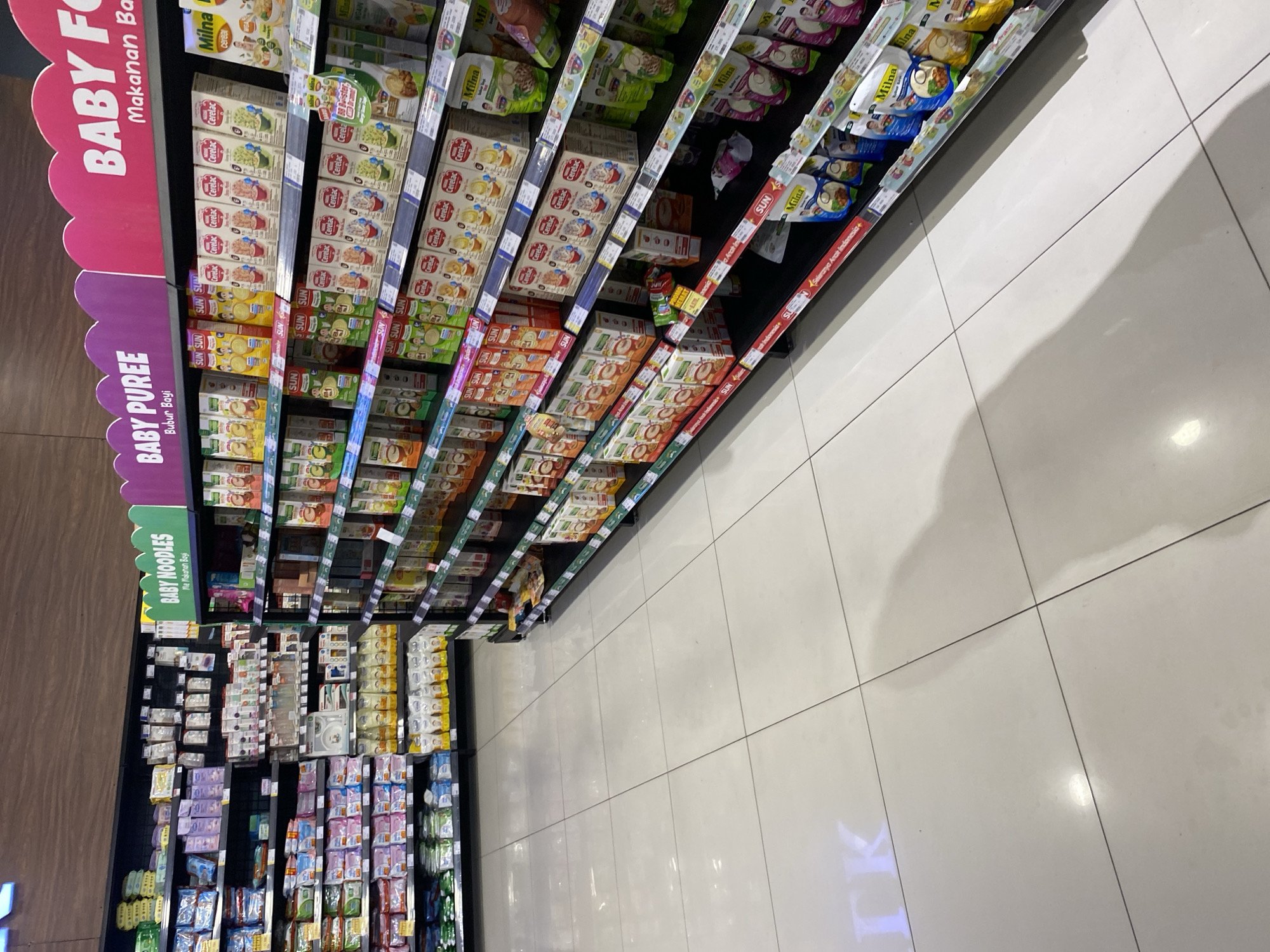 The image shows a wide aisle in a well-lit supermarket, specifically a baby food section. The shelves are fully stocked with various baby food products, including cereals, purees, and snacks, in colorful packaging. Brands like "Cerelac," "Sun," and "Milna" are prominently displayed. The products are arranged on multiple shelves, with some items in boxes and others in pouches. The flooring is a clean, light-colored tile with visible grout lines. The overall impression is of a modern and organized retail environment. Based on the presence of brands and common supermarket layouts, this could be a store in Banda Aceh, Indonesia, or any other city with similar retail infrastructure. No individuals are visible in the frame.