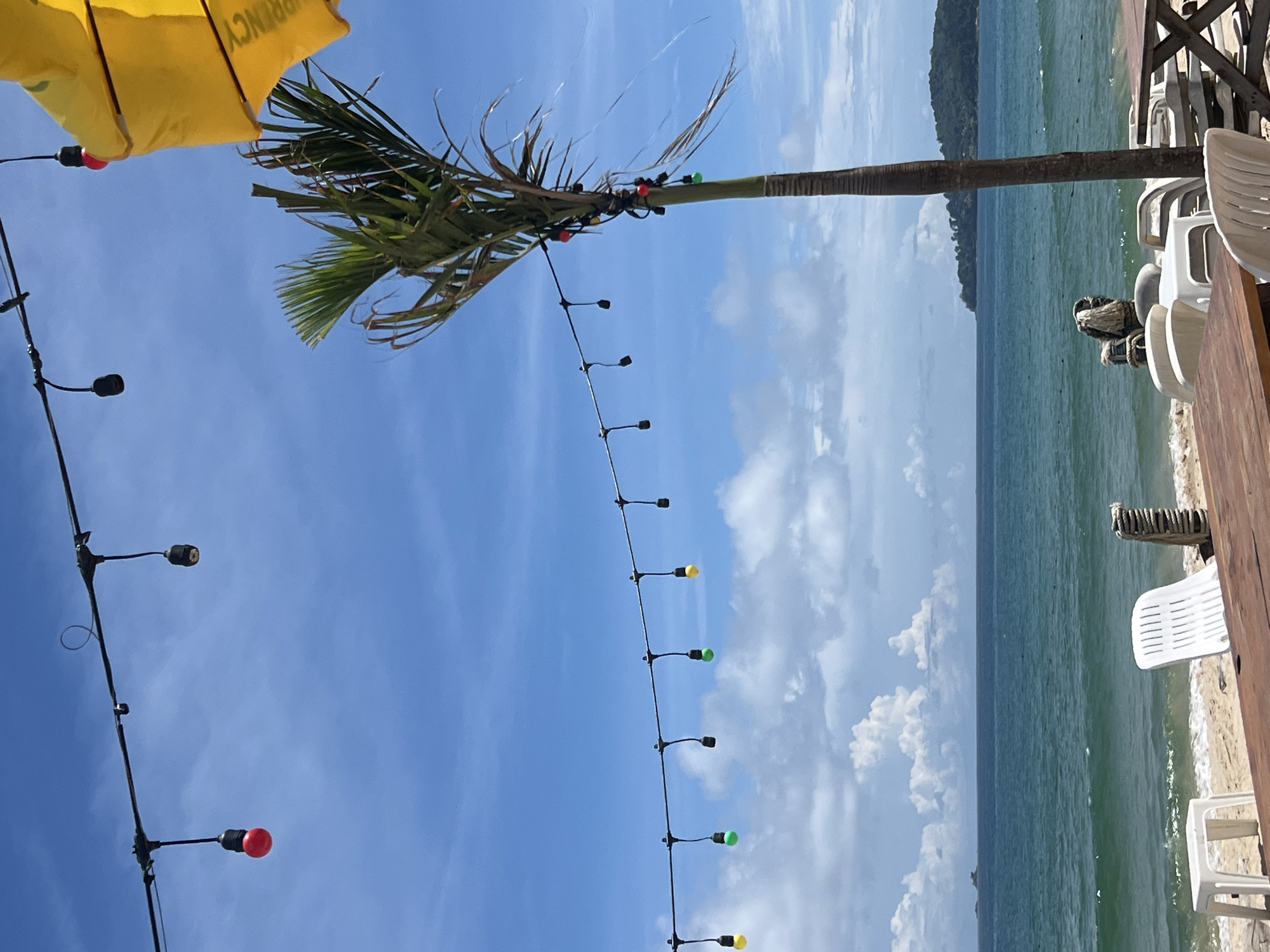 A string of colorful lights hangs across a beach scene in Choeng Thale, Thailand. The sky is blue with scattered white clouds. A tall palm tree stands on the right, adorned with some of the lights. The ocean is visible in the background, with a distant island on the horizon. White plastic chairs and wooden tables are arranged on the sandy beach, suggesting a cafe or restaurant setting. A yellow umbrella is partially visible in the upper right corner.