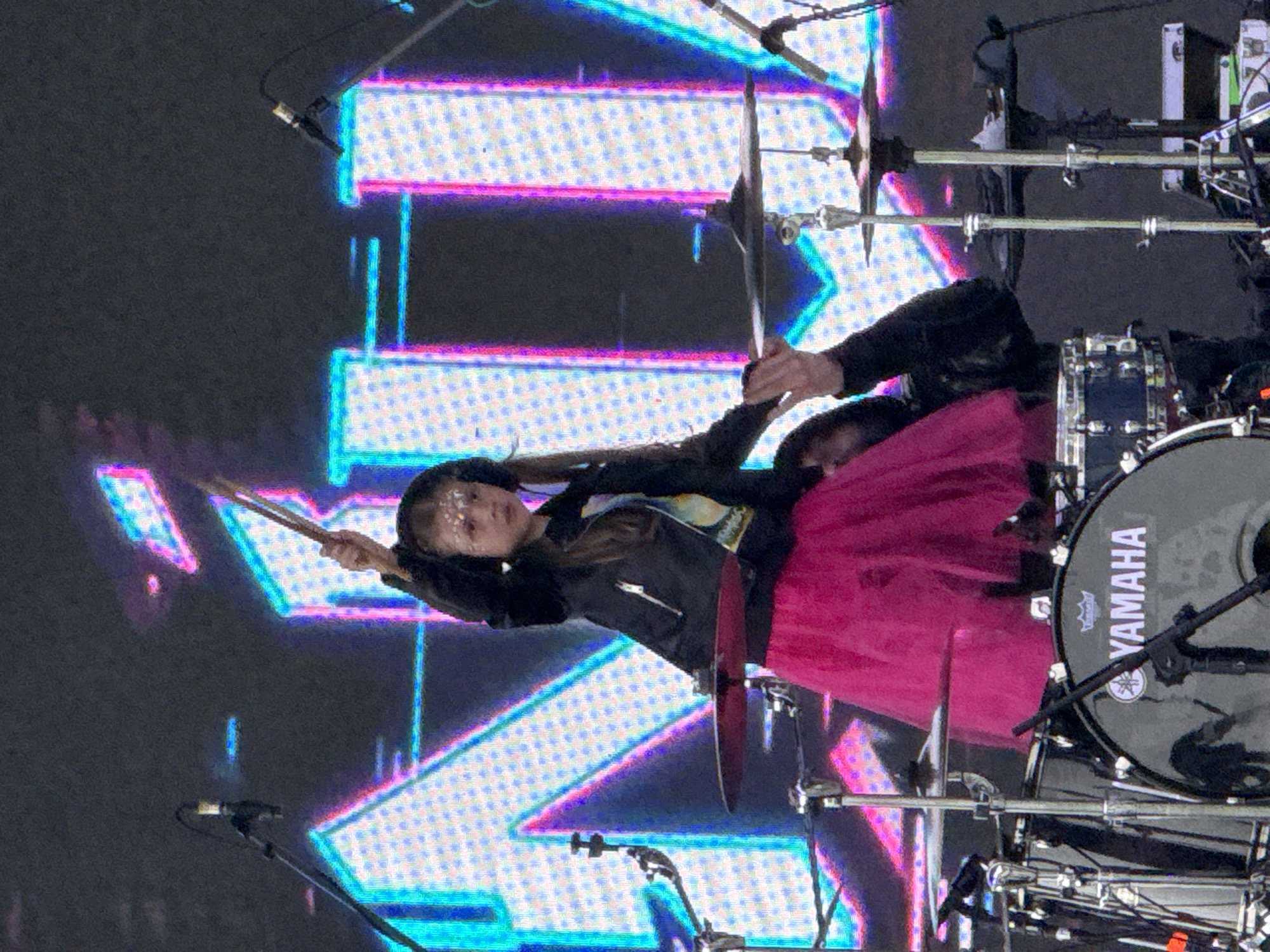 This image captures a live performance on a stage in Большая Кивара, Russia. The central figure is a young girl, estimated to be between 6 and 10 years old, with long brown hair, wearing black over-ear headphones, a black leather-style jacket, and a bright pink tutu-style skirt. She has reflective adornments applied to her forehead. The girl is positioned behind a drum kit, holding a pair of drumsticks high above her head with her right hand, poised to strike. Her left hand rests on a cymbal, overlaid by an adult's hand, presumably providing guidance or assistance.
Visible components of the drum kit include a black Yamaha bass drum, clearly branded with "YAMAHA" in white lettering, located in the lower left quadrant. Multiple cymbals are present, along with various drum stands and a partially visible blue snare drum. Several microphone stands are positioned around the drum kit. The background consists of a large LED screen displaying vibrant, pixelated, neon-colored geometric patterns, resembling abstract letters in shades of cyan, pink, and white against a dark backdrop. The overall scene suggests a musical performance in a stage environment.
