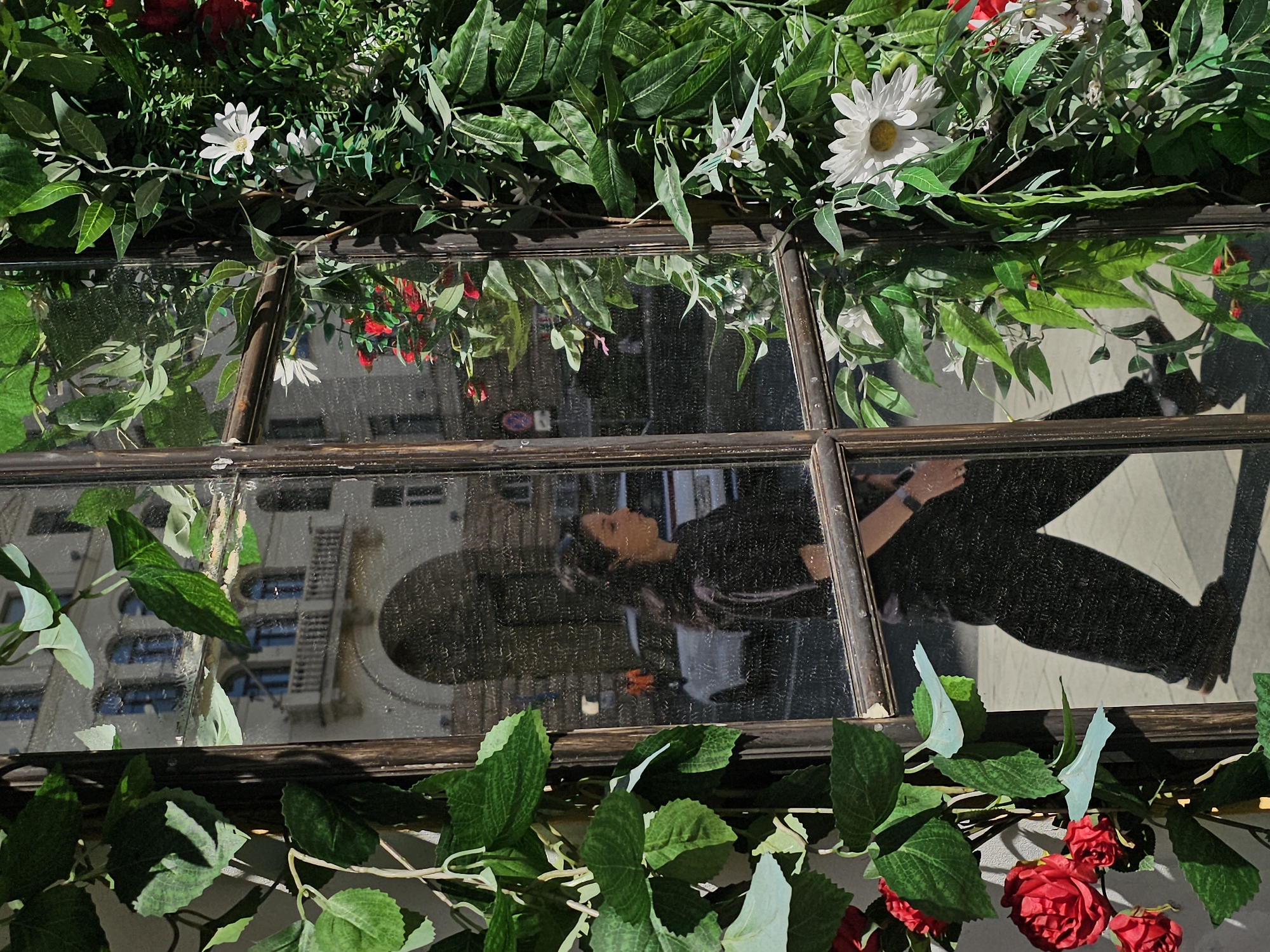 The image depicts a reflection in a window pane, framed by artificial greenery and red roses. The reflection shows a woman with dark hair, wearing a black outfit, walking on a sidewalk. Behind her, a white car is visible, as well as a person in an orange jacket. The background of the reflection reveals architectural elements, including an arched entryway and a building with balconies, suggesting an urban environment. Based on the architectural style, the location is likely the Arbat Street area in Moscow, Russia, known for its historic buildings and pedestrian-friendly atmosphere. The window itself has a rustic, wooden frame and appears to be part of a shop or cafe, adorned with lush green artificial foliage and artificial flowers, creating a decorative facade.