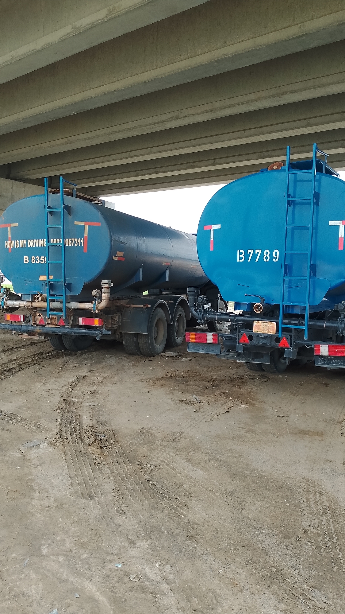 The image shows two blue tanker trucks parked in Effurun, Nigeria. The trucks are positioned beneath a concrete overpass. The ground is dirt with tire tracks. The first truck displays the text "HOW IS MY DRIVING 09032067311" and "B 8359." The second truck displays "B 7789" on its tank. Each truck has a ladder leading to the top of the tank and a red and white "T" logo.