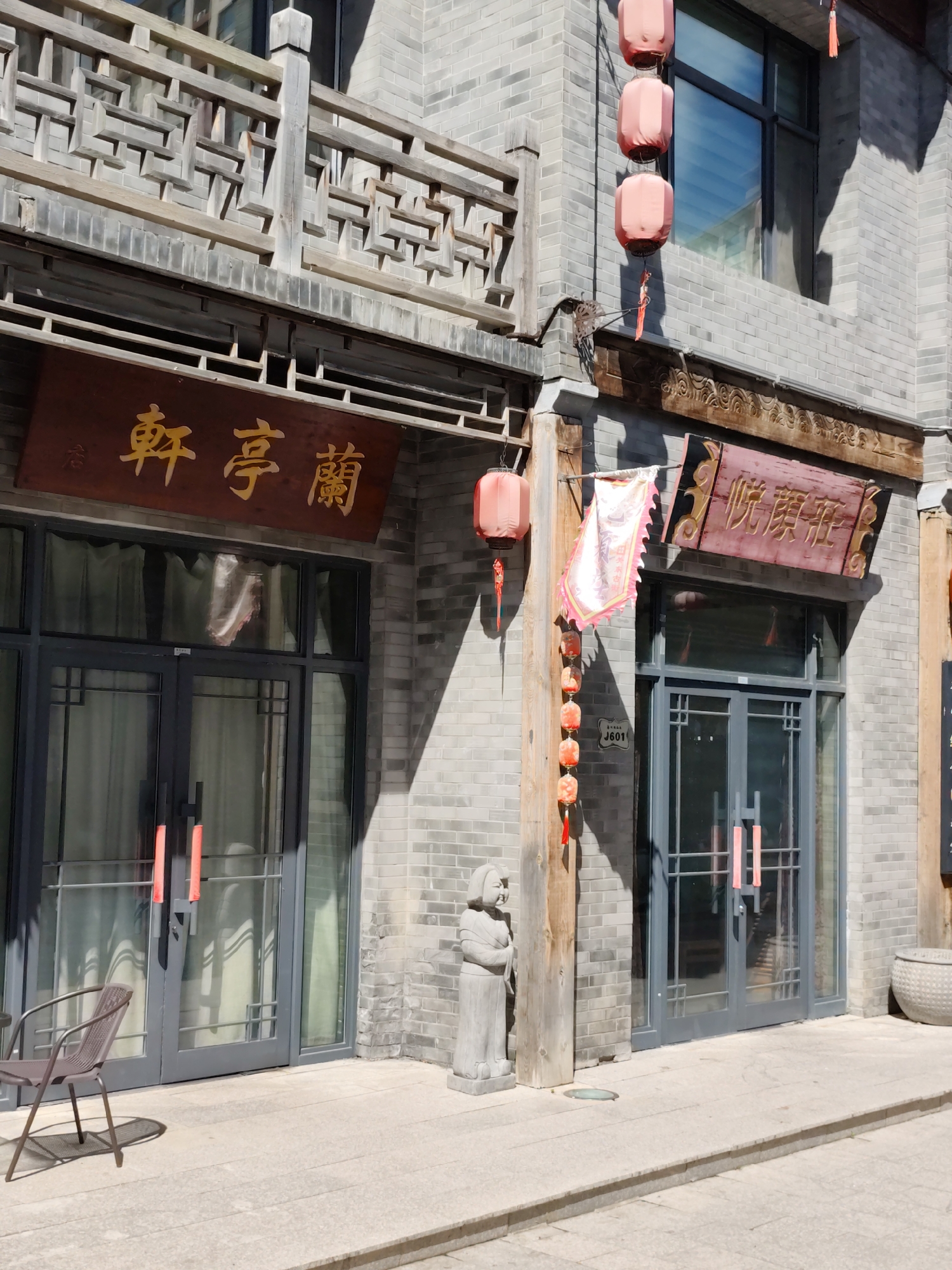 In Lindian, China, a traditional courtyard-style building features grey brick walls and wooden architectural elements. Red lanterns hang from the eaves, adding a splash of color to the scene. A dark brown wooden sign with golden Chinese characters is mounted above the entrance of the building, as are other wooden signs with Chinese lettering. A small statue of a person in traditional clothing stands on the paved walkway in front of the building. A modern metal chair sits on the pavement. The sun casts shadows from the architectural features onto the building and pavement.