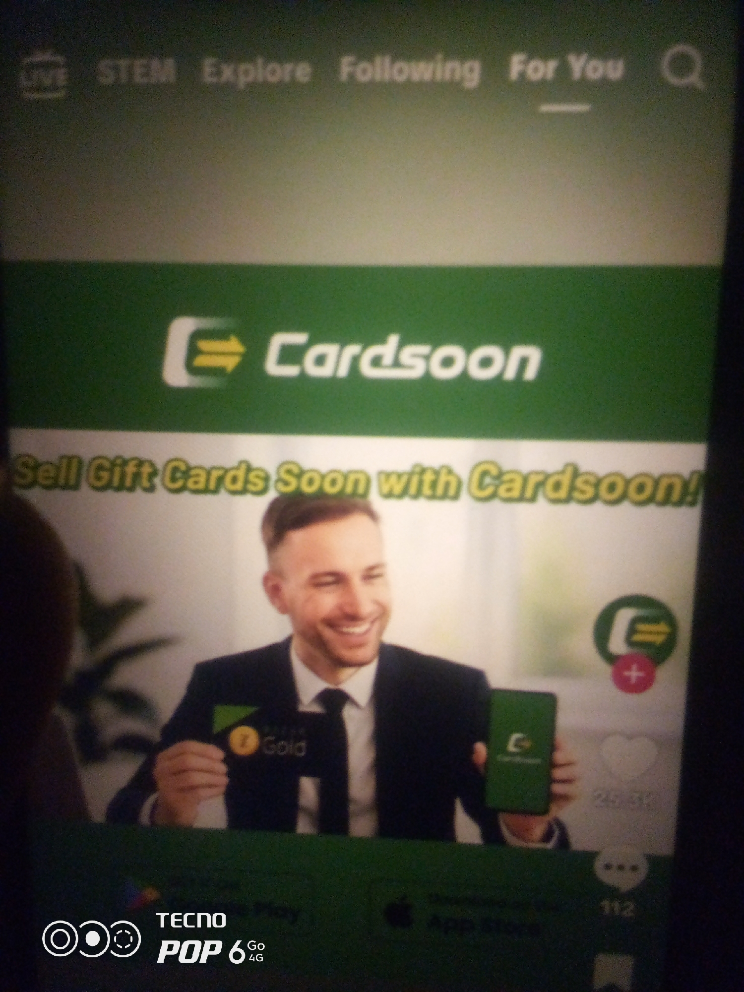 The image shows a screenshot of a mobile application, likely from a social media platform, featuring an advertisement for "Cardsoon". The ad prominently displays the "Cardsoon" logo, a green rectangle with two yellow arrows forming an "E", and the tagline "Sell Gift Cards Soon with Cardsoon!". A smiling man in a suit and tie is shown holding a phone displaying the Cardsoon app and a gift card labeled "7 Gold". To the right of the man, there are social media interaction icons, including a "like" button with a heart and a number indicating engagement. At the bottom of the screen, the "TECNO POP 6 4G" logo is visible, suggesting the device on which the screenshot was taken. The background is blurred and appears to be a light gray or off-white color, with a dark shadow on the right side of the image. The overall scene is an advertisement for a digital service. There is no indication of the location being Howayi, Nigeria.