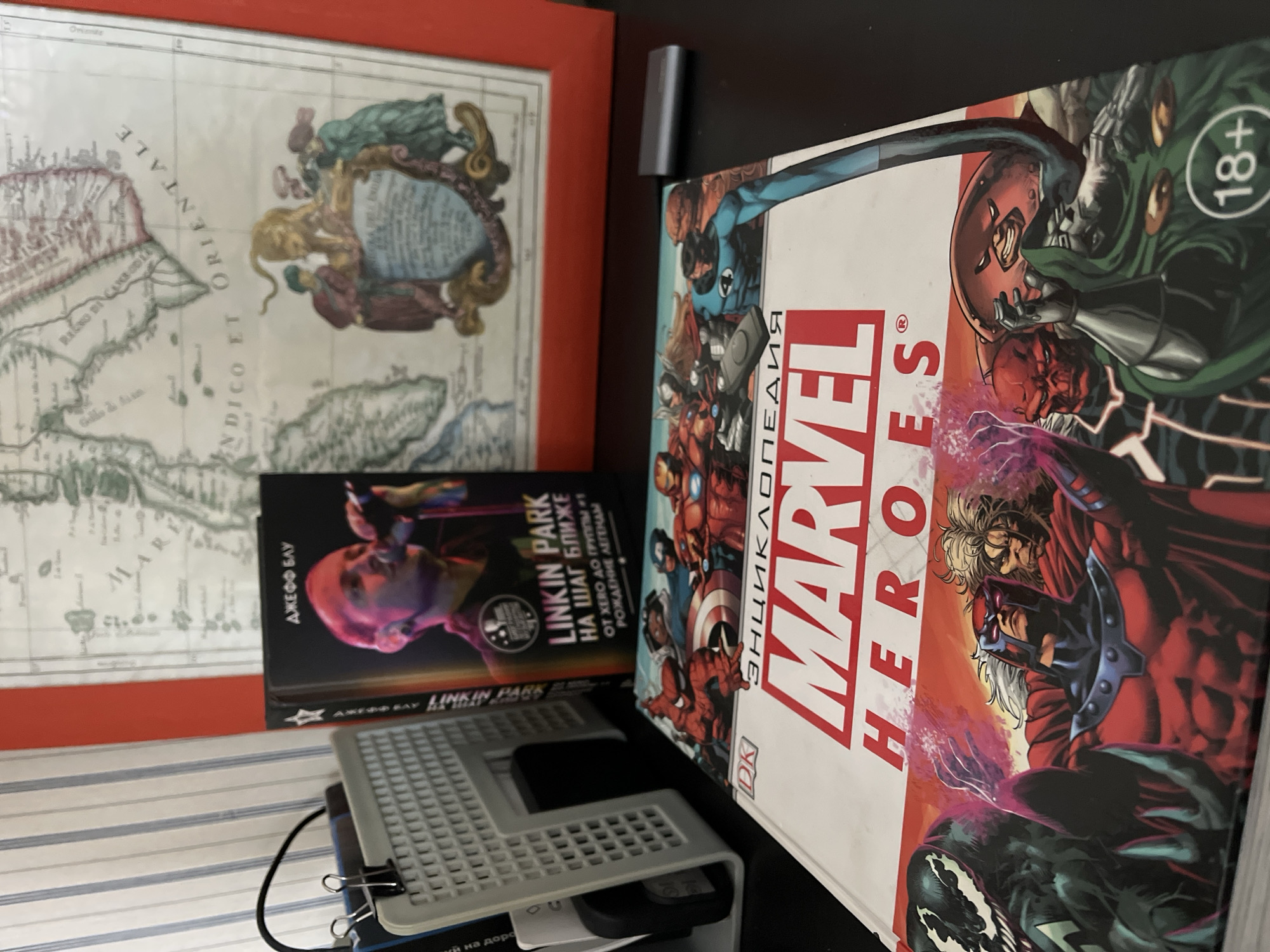 The image shows a collection of items stacked on a surface. On the left, a framed map with a red border is visible. In front of the map, there's a book titled "Linkin Park" by ДЖЕФФ БАУ in Russian. Below the book is a white computer keyboard stacked on top of other items including a smartphone, secured by a black clip. To the right, a thick book titled "MARVEL HEROES" in Russian is prominently displayed, featuring comic book characters on its cover. An "18+" sign is visible in the upper right corner of the Marvel book. The overall environment appears to be indoors. It is impossible to determine the location as Krasnoyarsk, Russia from the image alone.