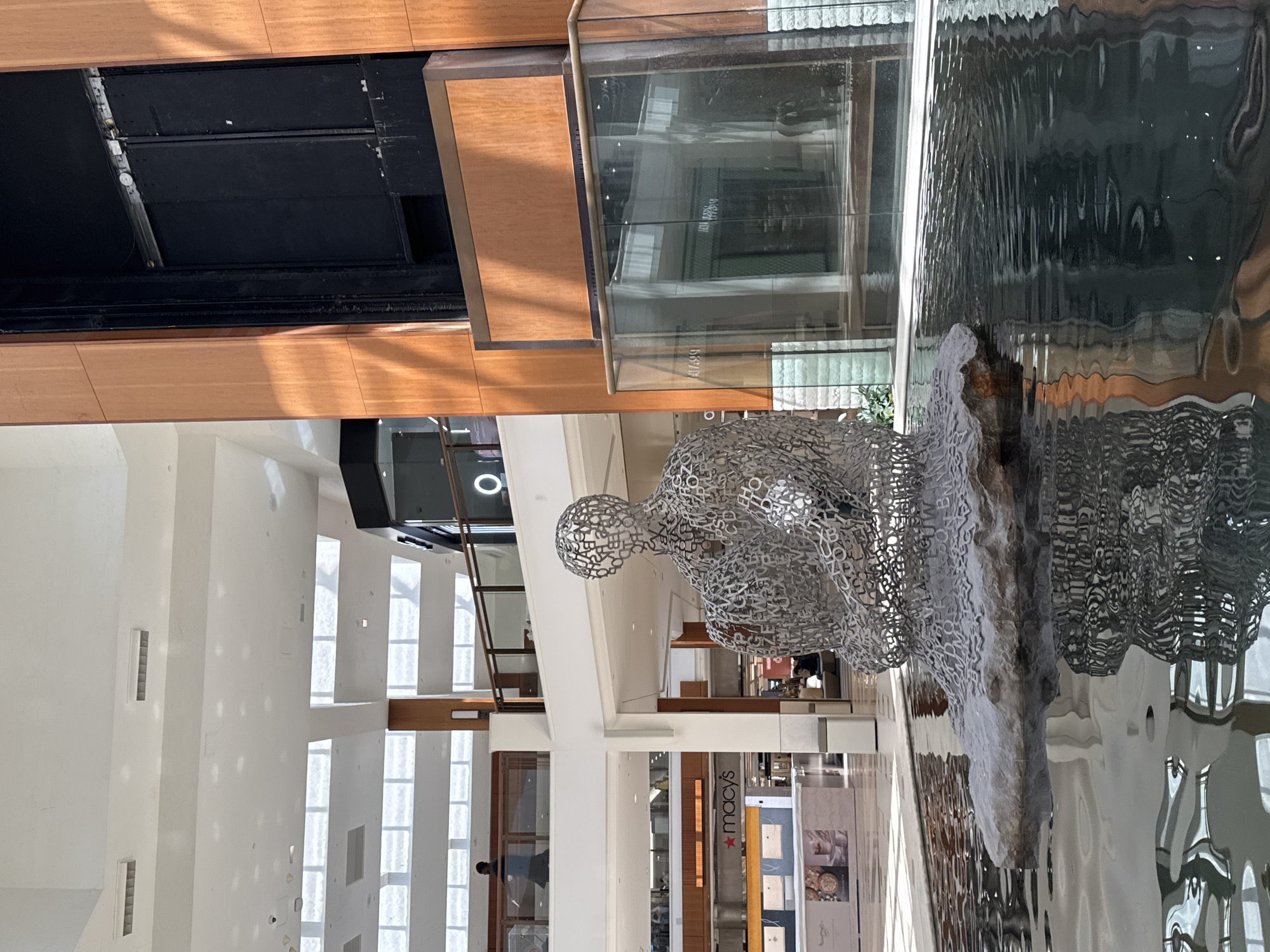 The image depicts an interior scene within the Aventura Mall, located in Aventura, Florida, USA. A sculpture of a seated figure, constructed of a metal framework forming letter shapes, is a primary subject. The sculpture is near a water feature. The scene is situated within a shopping mall environment. In the background, shop fronts can be seen, including a Macy's store. The overall impression is one of a modern architectural space integrating art and commercial activity. Natural light streams into the space through multiple windows along a wall.