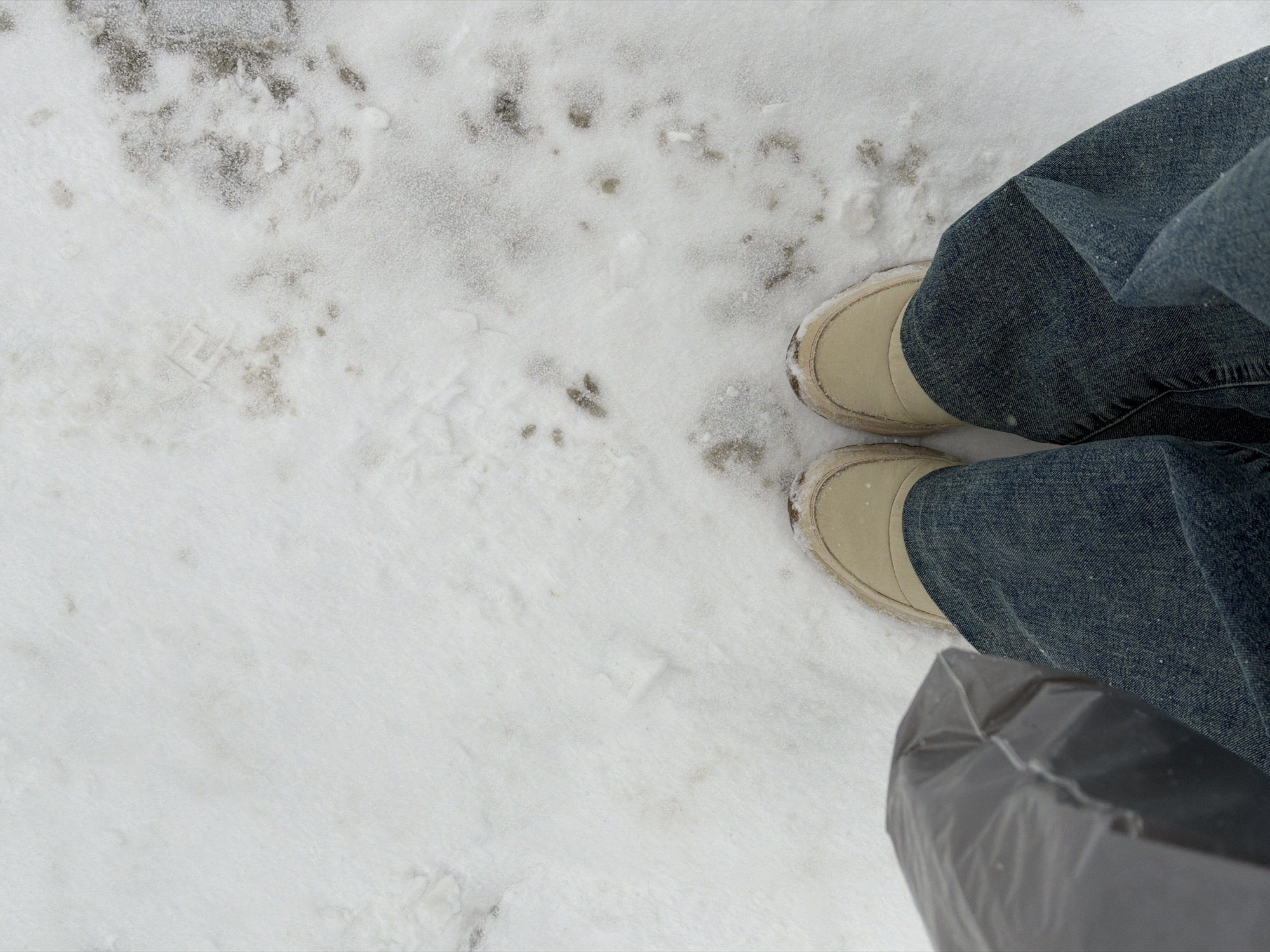 The image shows a view from above of a person standing in snow, likely on a street. The person is wearing blue jeans and tan-colored winter boots. The ground is covered in a mix of fresh snow and areas that have been dirtied, likely by traffic or foot traffic. A black plastic bag is also visible near the bottom of the image, possibly being carried by the person. The location might be Berezniki, Russia, implied by the user's description.