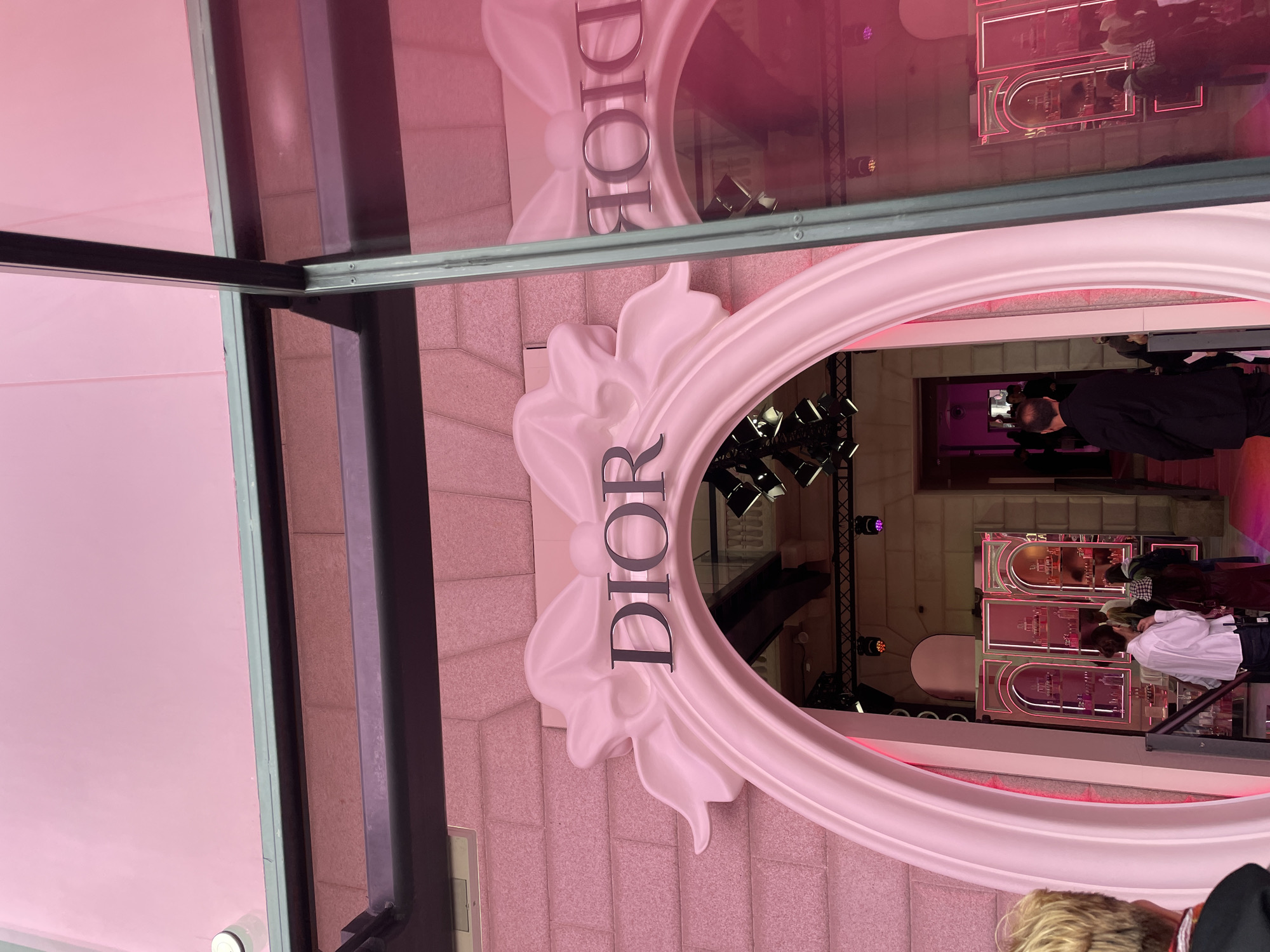 The image shows a view of a Dior store in Milan, Italy, seen through a window. The Dior logo, displayed in silver lettering within a large, ornate white frame, is prominently visible on the exterior of the building. The reflection in the window shows a reversed image of the logo and some elements of the interior. Inside the store, lighting equipment is visible, along with shelves displaying products with pink lighting. People are present inside the store, including individuals wearing black jackets and white shirts. The exterior wall of the store is textured with a pink hue. The window frame is black.