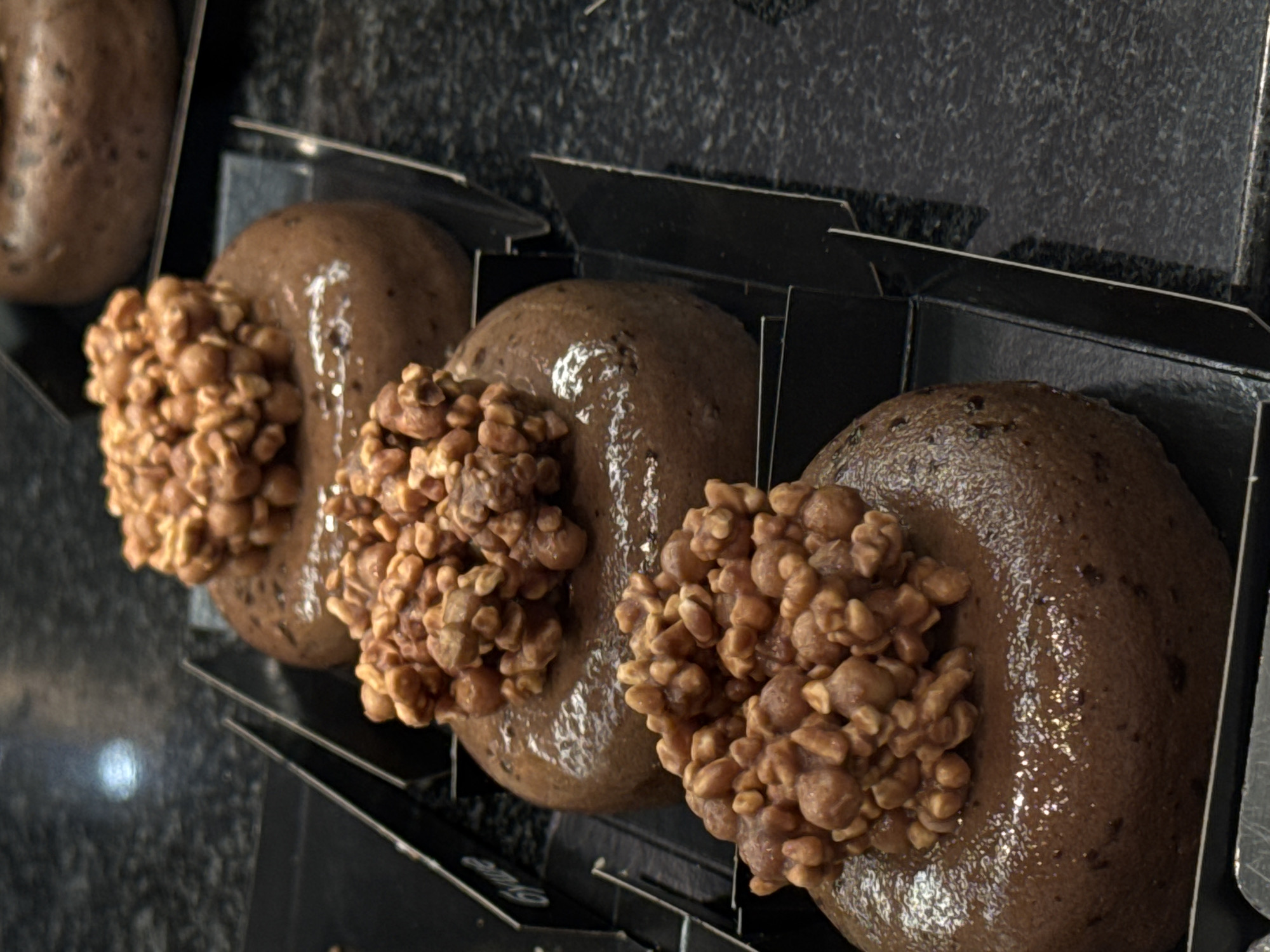 The image shows a close-up of three donuts in individual black boxes. The donuts are round, covered in a smooth, dark brown glaze, and topped with a cluster of light brown, nut-like pieces. They are arranged in a row from left to right. The background appears to be a dark, possibly granite or marble, surface. The location is Saint Petersburg, Russia.