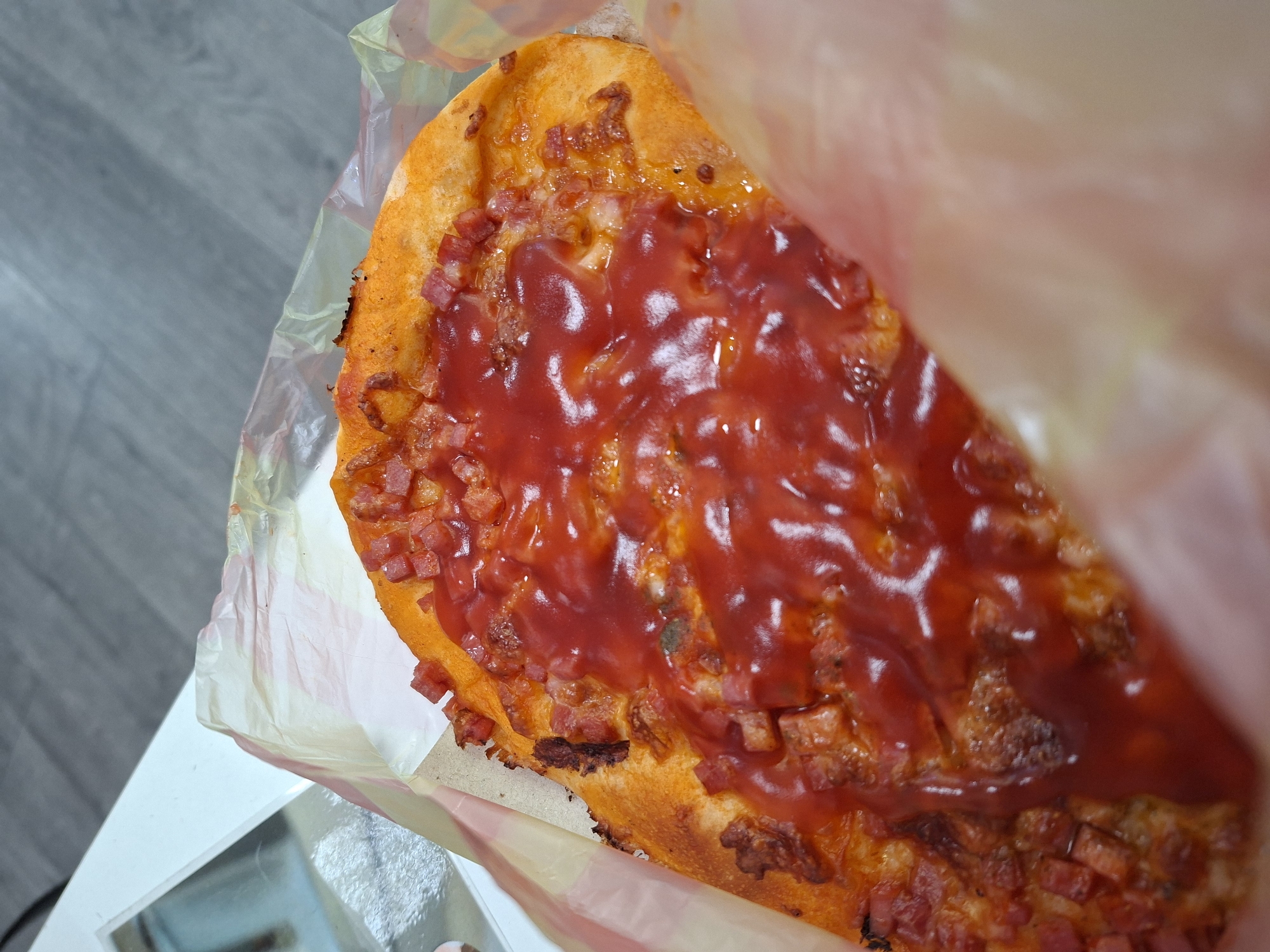 A partially eaten pizza slice is presented inside a partially open plastic takeout bag on a white table with a reflective surface. The pizza features a red sauce covering a topping of small, cubed pieces of a cured meat product. The crust appears to be slightly burnt along the edge. The table sits on a gray wood-patterned floor. It is unclear if this is Galabovo, Bulgaria.