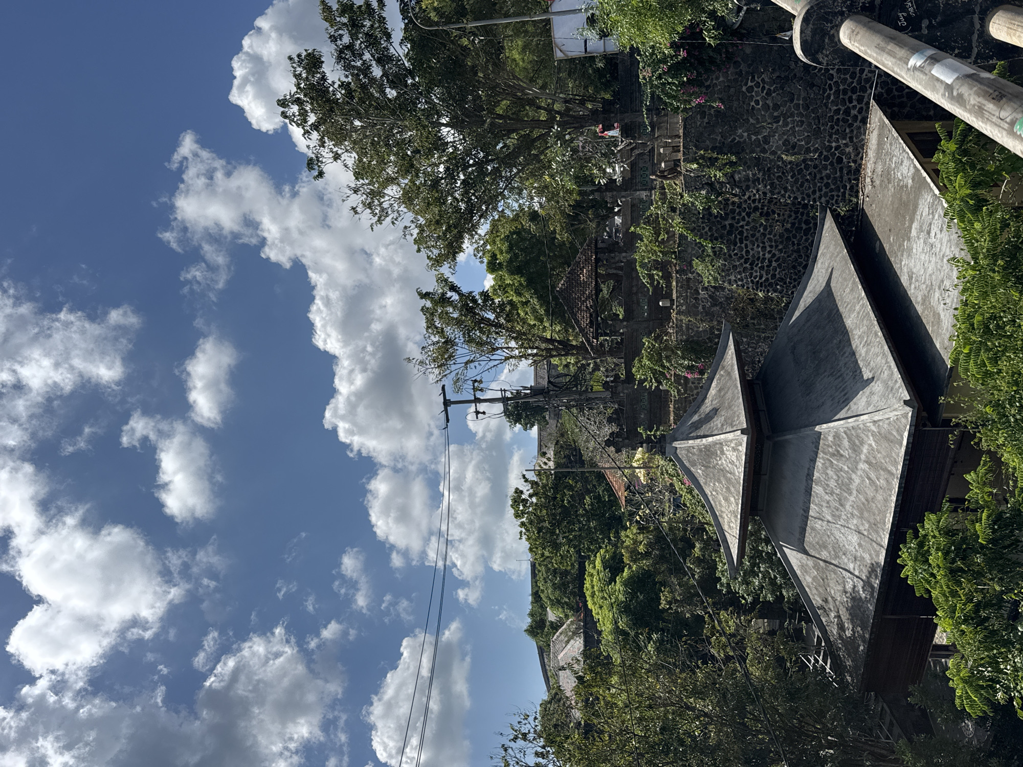 The image depicts a multi-tiered building with a gray, pagoda-style roof, nestled amongst lush green trees and a stone wall. The building's architecture suggests a tropical or Southeast Asian setting. In the background, more buildings with tiled roofs and abundant vegetation can be seen on a hillside. A clear blue sky with scattered white clouds dominates the upper portion of the frame. Utility poles and wires are visible, indicating the presence of modern infrastructure within the natural and architectural landscape. The scene is likely a public space or a religious site, given the architectural style and the serene natural surroundings. The location is identified as Kuta Selatan, Indonesia, a region known for its scenic beauty and cultural heritage.