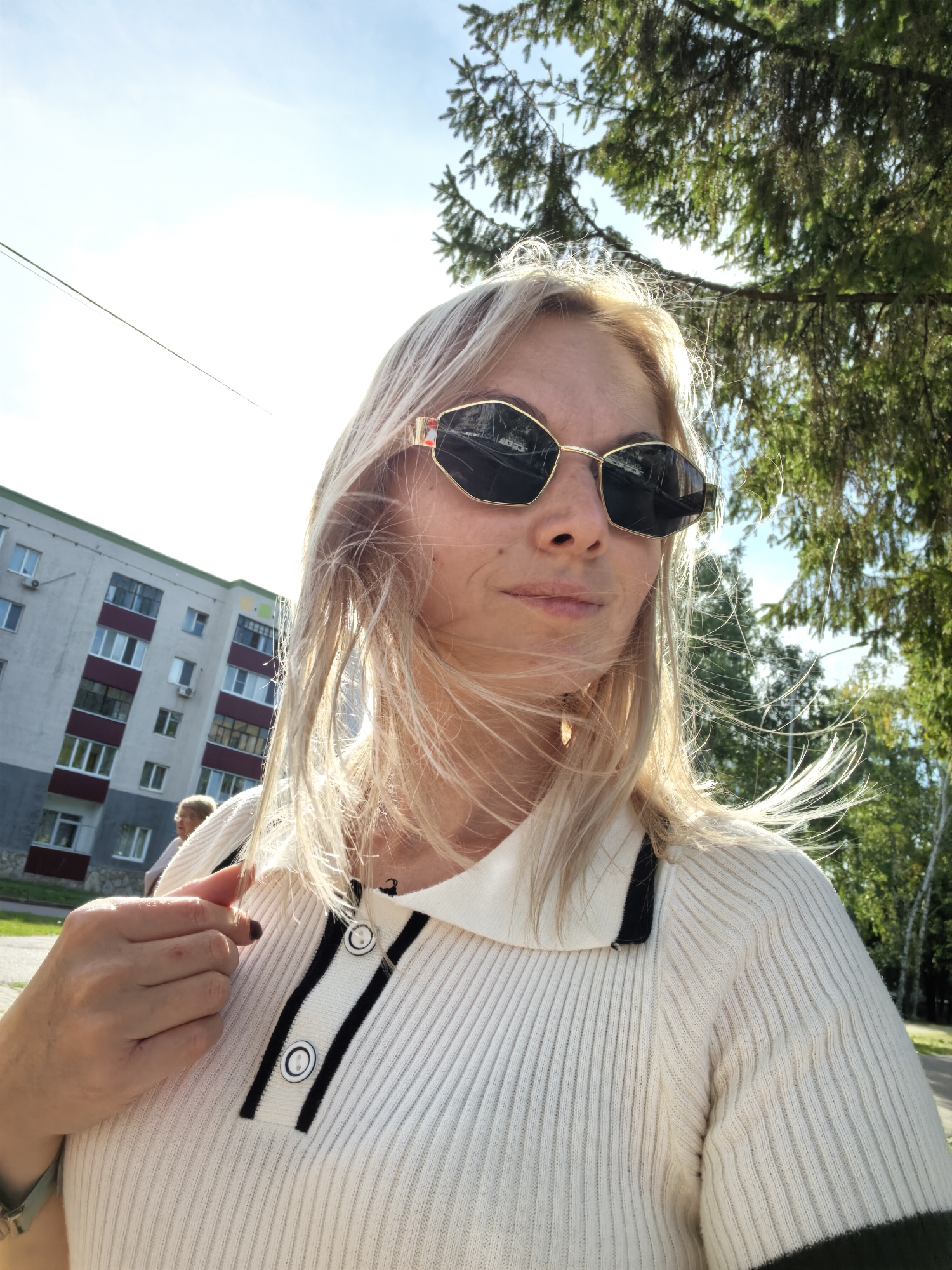 A woman with blonde hair and sunglasses is taking a selfie outdoors. She is wearing a white ribbed sweater with black and white trim and buttons. In the background, there is a multistory apartment building and a tree with green foliage. The sky is blue with some clouds. The image appears to be taken during the daytime.