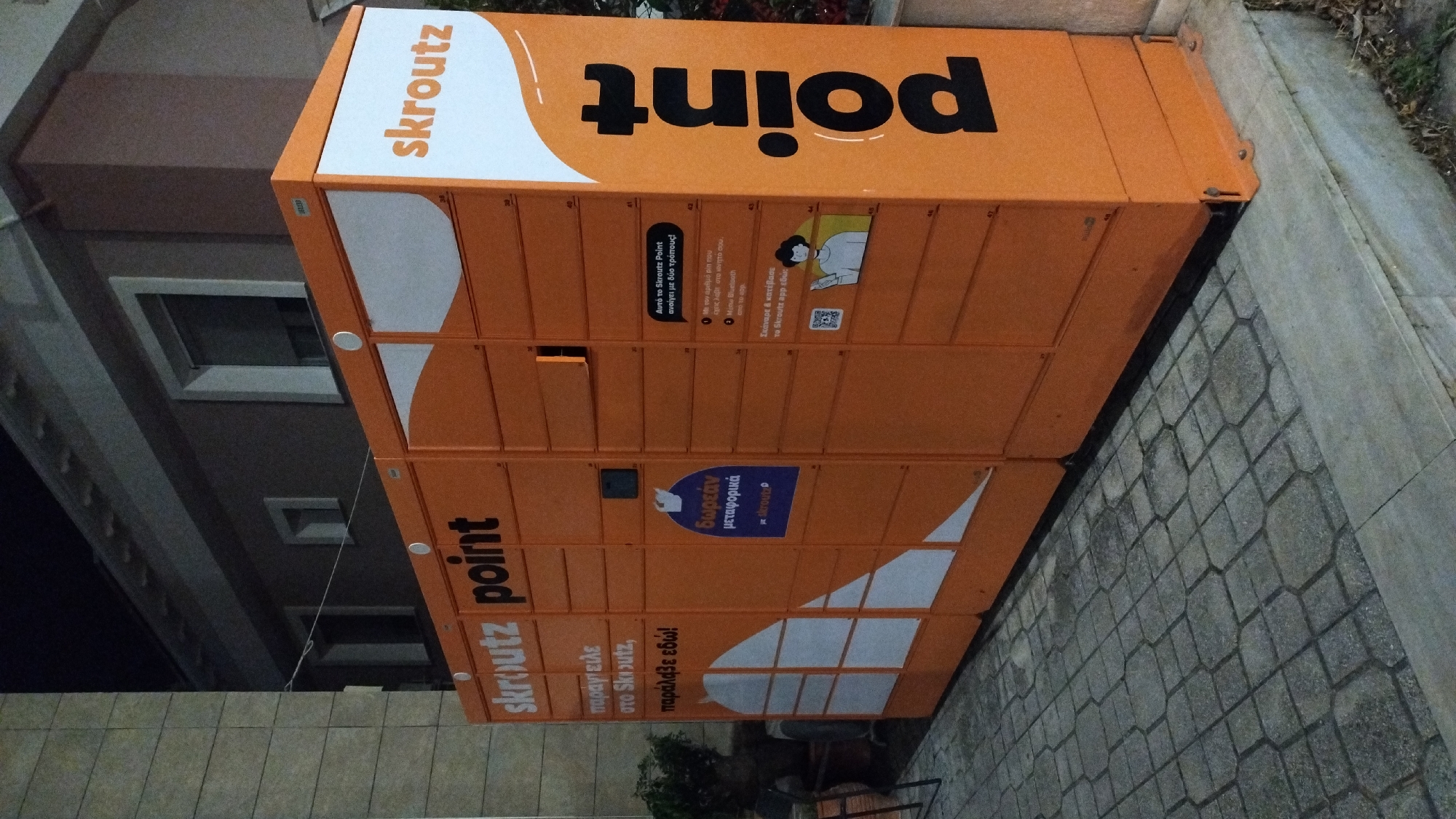 This media file captures an orange parcel locker, branded with "skroutz point," situated outdoors. The locker is a self-service delivery point, likely in Greece given the Greek text on its surface. The prominent text on the top reads "point" in large black lettering, and "skroutz" in orange to its left. The locker itself is divided into multiple compartments, suggesting it's designed for package retrieval and drop-off.
Below the "point" text, there's an informational panel with instructions in Greek. One section explains that the locker can be opened in two ways: via a code received on a mobile phone or through Bluetooth from the Skroutz app. Another blue panel highlights "δωρεάν μεταφορικά," meaning "free shipping." Further down, more Greek text appears, reading "παράγγειλε στο Skroutz, παράλαβε εδώ!", which translates to "order from Skroutz, receive here!".
The setting appears to be a street or sidewalk, with cobblestone paving visible in the foreground and a textured wall or building in the background. The lighting suggests it might be evening or nighttime, as the surroundings are somewhat dimly lit, although the locker itself is well-illuminated. The overall impression is of a functional, modern piece of urban infrastructure facilitating online shopping logistics.