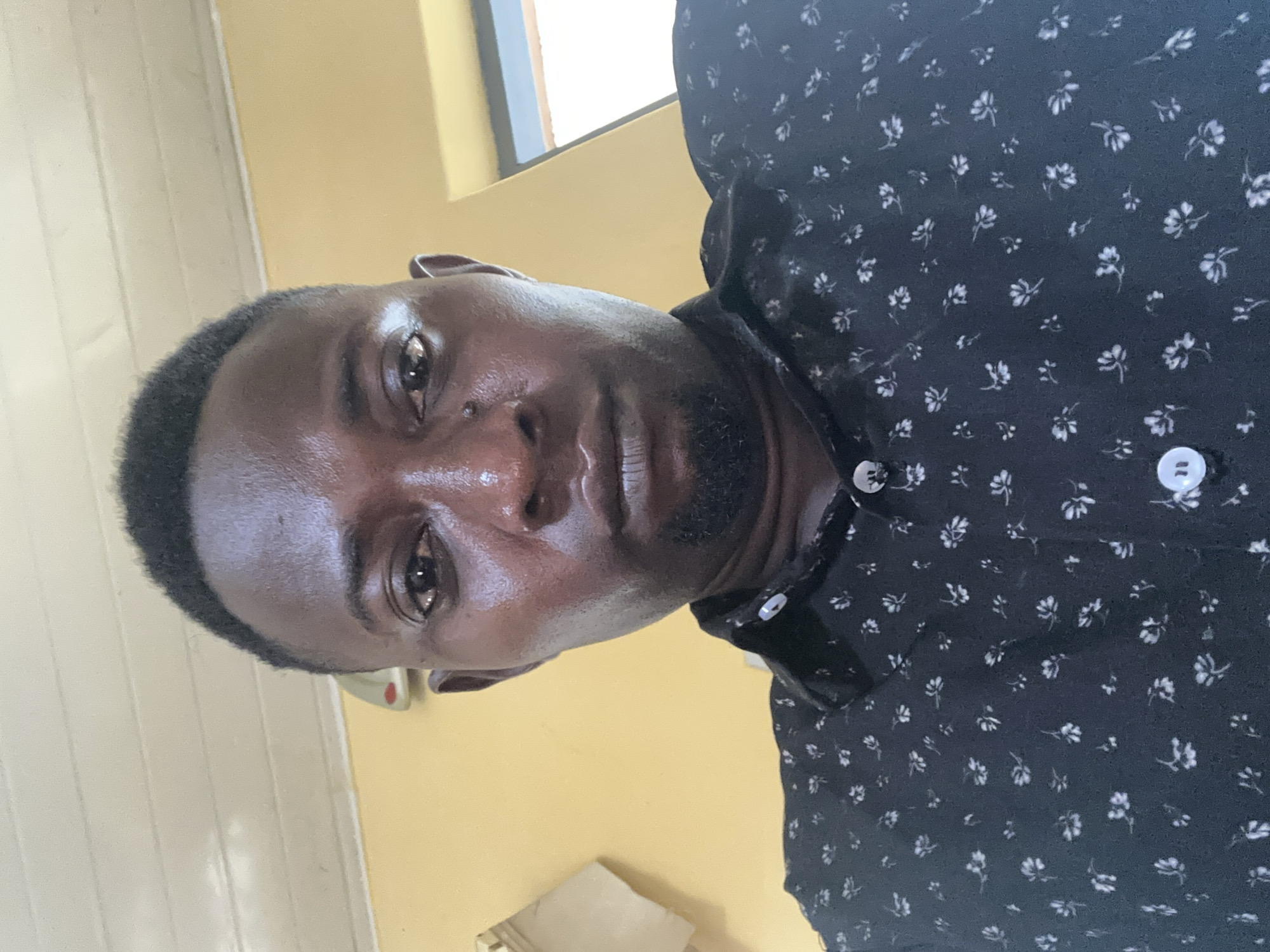 The image is a close-up, low-angle selfie of a Black man in Kaduna, Nigeria. He has dark skin, short dark hair, and is wearing a dark collared shirt with a white floral pattern. His expression is neutral. The background appears to be an indoor setting with a yellow wall and a white ceiling with horizontal panels. A window with a view of what appears to be an exterior structure is visible on the right side of the frame. There is also a light-colored object, possibly a shelf or a piece of furniture, on the left side. The lighting suggests it is daytime.