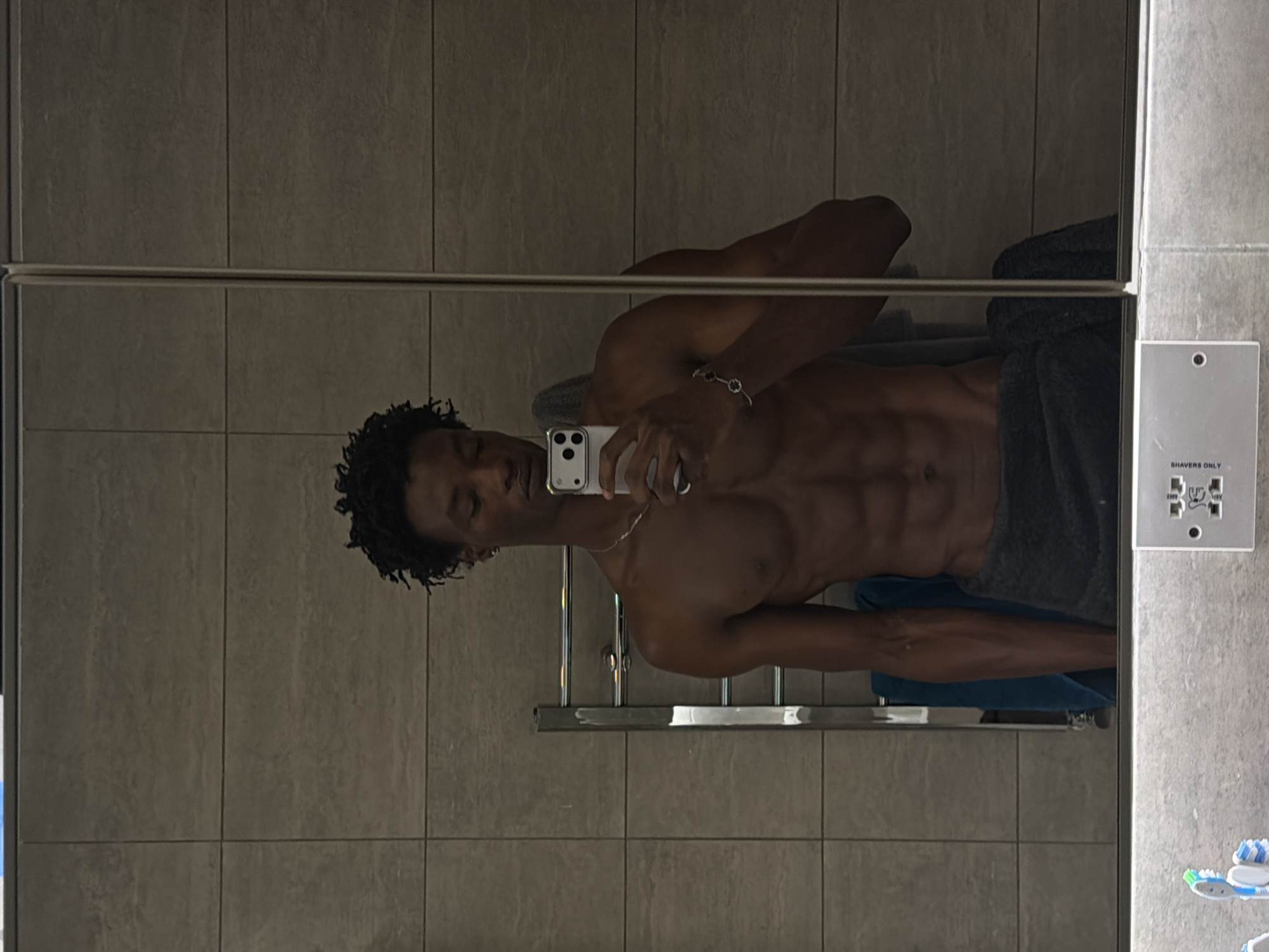 A shirtless man with dark skin and curly hair is taking a mirror selfie in a bathroom. He is wearing a dark grey towel around his waist. The bathroom has tiled walls and a towel warmer to his left. In the foreground, a shelf with toothbrushes and an electrical outlet is visible. The scene appears to be in a private residence, likely a bathroom. There is no visible indication of the city or country.