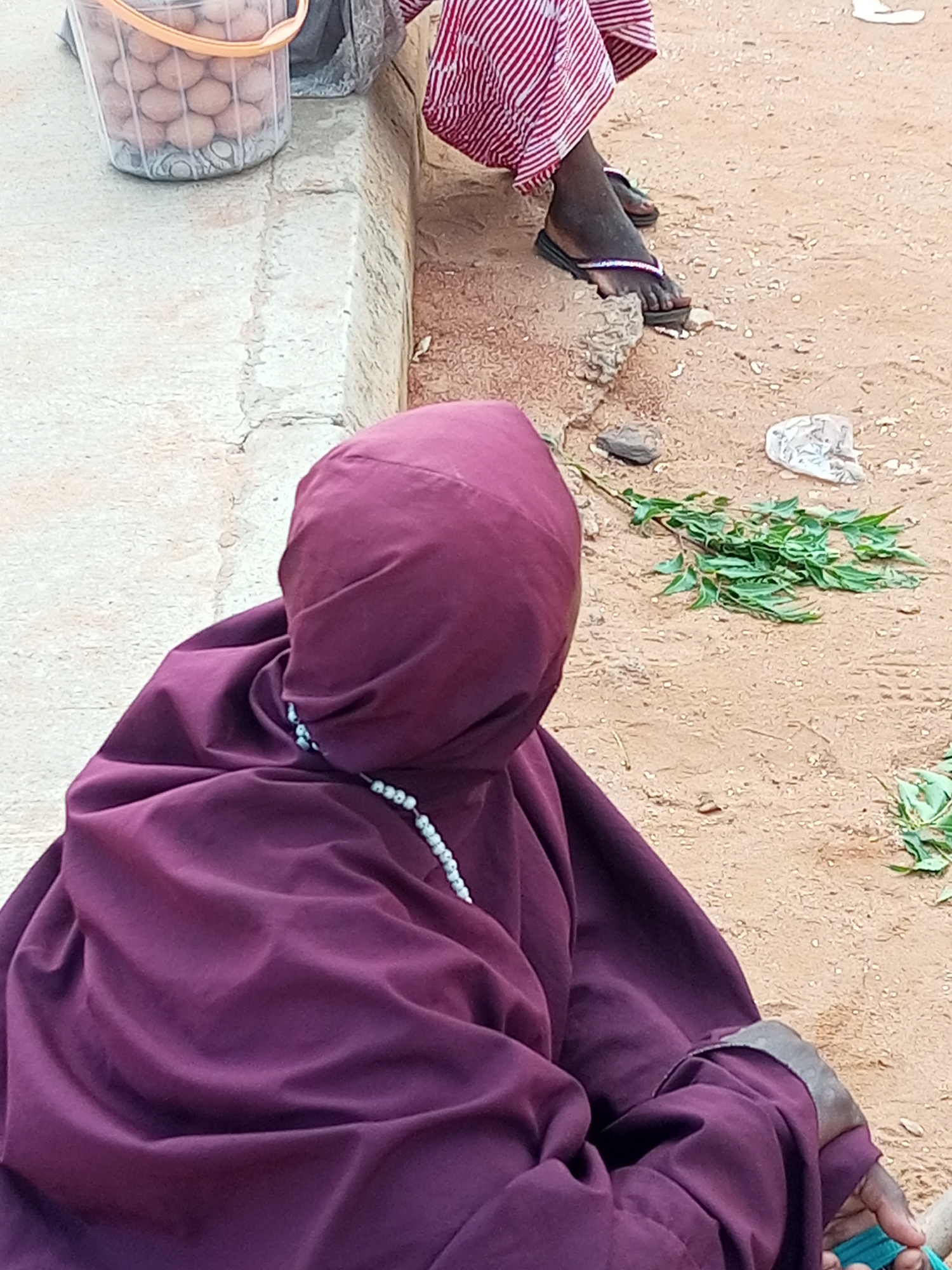 In Gombe, Nigeria, a person wearing a dark purple hijab and robe sits on the ground. A strand of white beads is visible around their neck, partially obscured by the hijab. The individual's hands are clasped in their lap. To the left of the person, a clear plastic bucket filled with eggs is placed on a concrete ledge. A second person, partially visible, sits to the right, wearing a red and white striped skirt and black flip-flops. The ground is sandy and dusty, with scattered green leaves and a crumpled piece of white plastic. The scene appears to be outdoors, with a weathered concrete structure forming a backdrop.