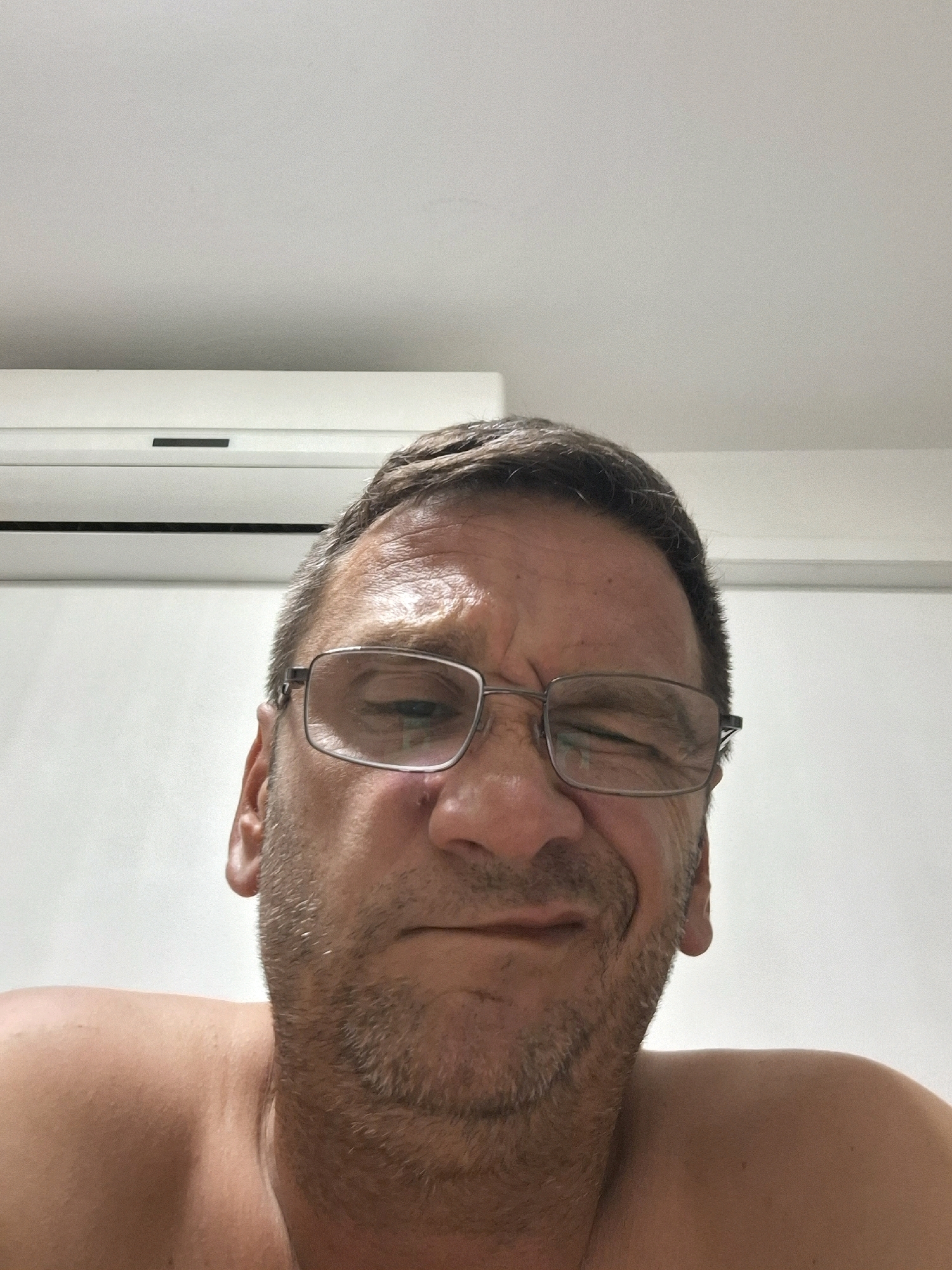 A close-up, selfie-style shot captures a middle-aged Caucasian male from the chest up, indoors, with an air conditioning unit visible in the background. The man is wearing eyeglasses and has short, greying hair and stubble. His expression is one of slight grimace or squinting. The background consists of a plain white wall and ceiling, with the air conditioning unit horizontally mounted. The scene appears to be a residential interior, possibly a bedroom, in Yerocham, Israel.