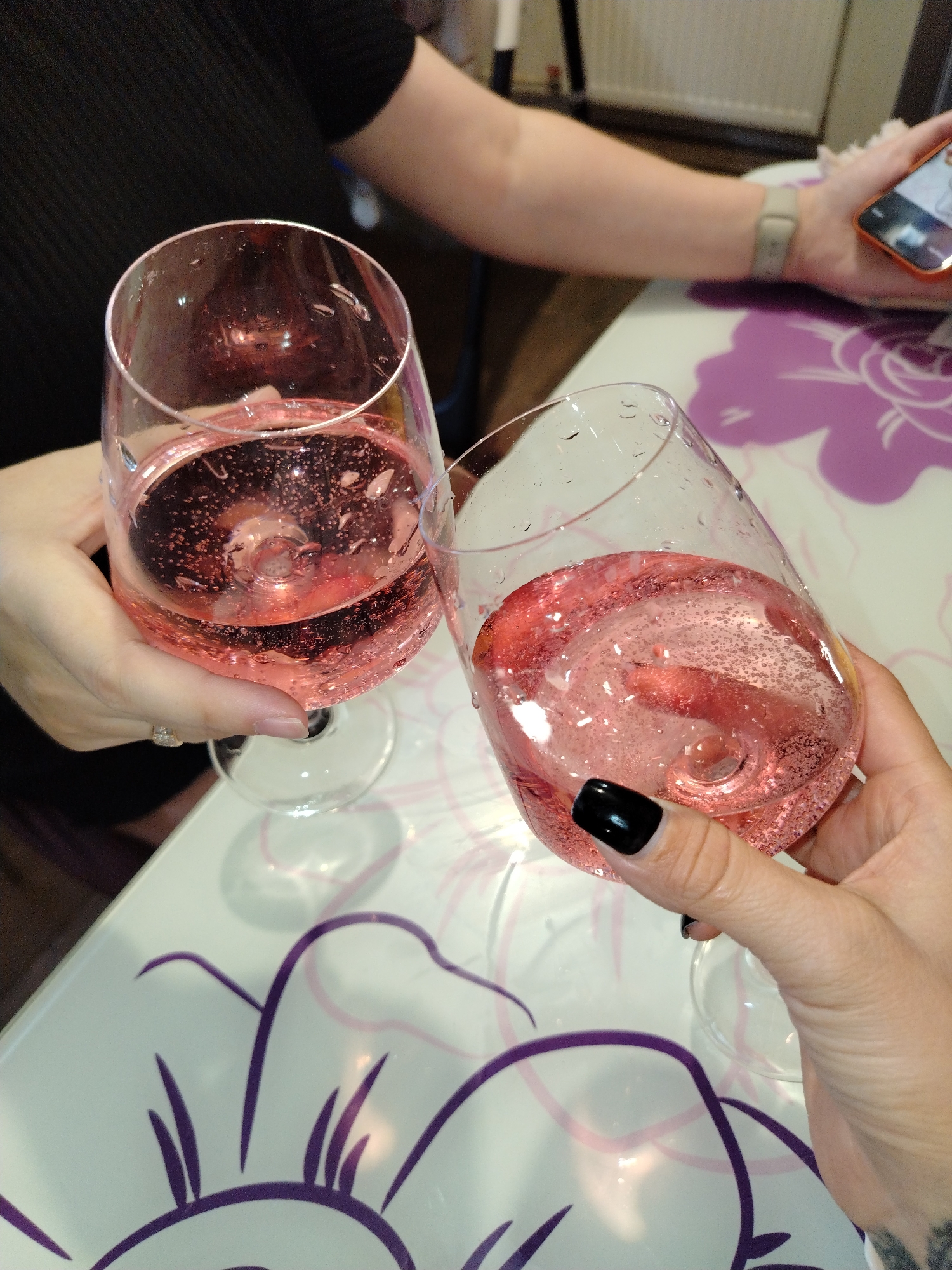 Two people are toasting with glasses of pink sparkling wine. One person holds a glass in their right hand, with black nail polish, and the other in their left hand, wearing a ring. A phone is visible on the table. The location is likely indoors, with a table featuring a white surface with purple floral designs. The city of Noviy, Russia, is not directly discernable from the image.