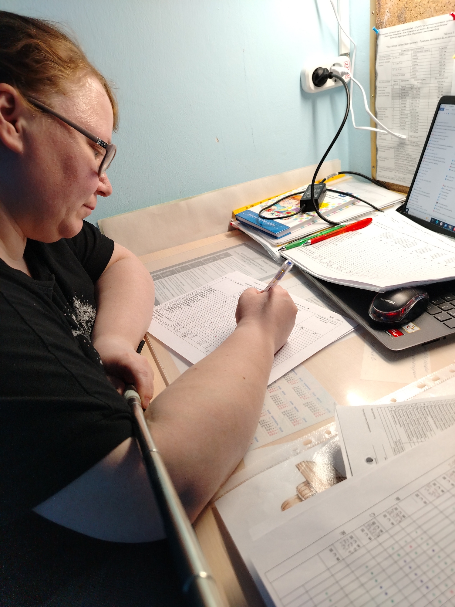 An adult woman with light brown hair and glasses is seated at a desk, focused on writing on a document. She is seen in profile from the chest up, wearing a black short-sleeved t-shirt with a light-colored design. Her left arm, extended into the foreground, holds a silver-colored object resembling a selfie stick or monopod.
The desk is cluttered with papers, documents, and various office supplies. The woman is actively writing with a blue pen on a form that features a grid and Cyrillic text. To her right, an open black laptop is visible, displaying what appears to be a spreadsheet or webpage. A black and red computer mouse rests on the laptop's keyboard. Several other pens (red and green) and a blue folder are stacked nearby. A printed calendar with Cyrillic days of the week ("Пн Вт Ср Чт Пт Сб Вс") is partially visible beneath other papers, along with more documents containing Cyrillic text and numerical data.
The background features a light blue wall. A white wall-mounted power outlet with multiple plugs connected is prominent, labeled "220V", confirming a European electrical standard. Cables run from the outlet to various devices on the desk. Above the laptop, a corkboard or similar notice board with a paper attached, also bearing Cyrillic text, is visible. The scene is illuminated by an artificial light source, casting a warm glow, suggesting it could be evening or night.
The woman appears concentrated on her task, engaging in an activity that involves both physical documentation and digital work. The visible Cyrillic text on the documents, calendar, and corkboard, combined with the "220V" electrical outlet, strongly indicates a setting in a Slavic-speaking country, consistent with the provided location context of Chaikovsky, Russia.