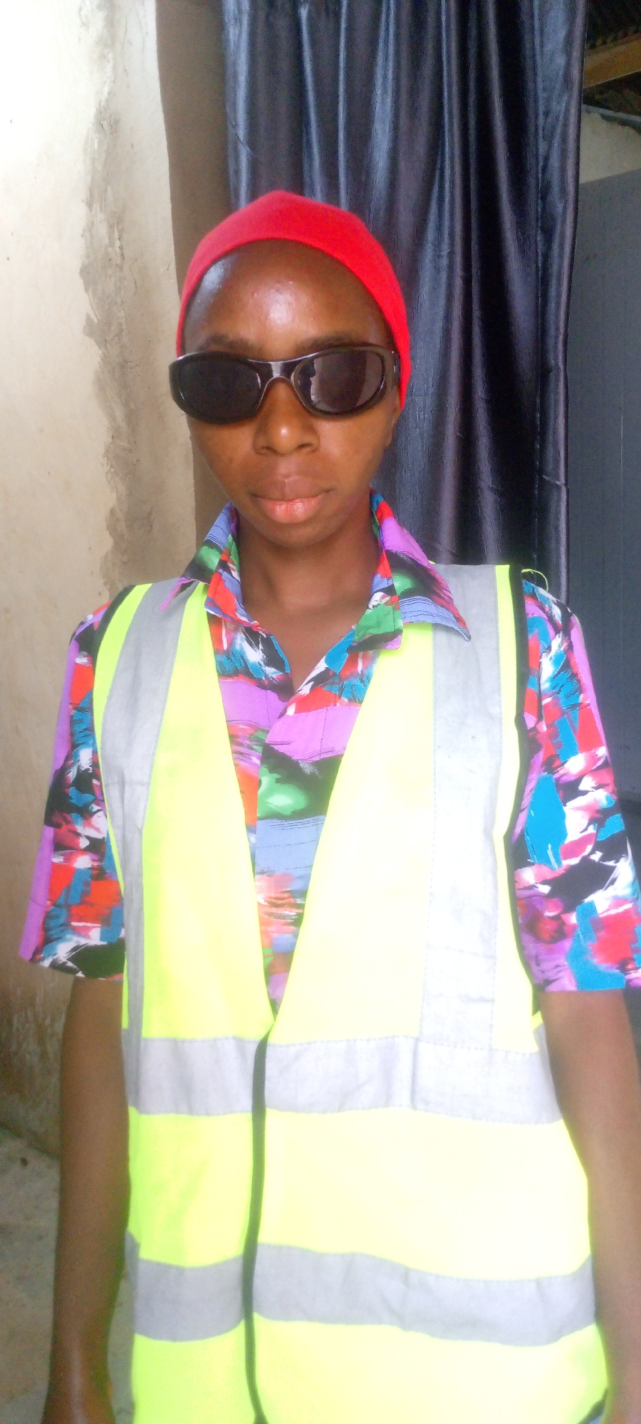 A young Black person, appearing to be a teenager or young adult, stands facing the camera outdoors. They are wearing a red bandana on their head and dark sunglasses. Their shirt is a vibrant, multi-colored pattern of abstract shapes and lines in pink, purple, blue, green, and black. Over the shirt, they wear a bright yellow safety vest with reflective silver stripes across the chest and torso. The background consists of a textured, light-colored wall on the left and a dark, draped material, possibly a curtain or tarp, on the right. The ground is not visible. The lighting suggests it is daytime. The scene is likely in a residential or casual outdoor area within a city in Nigeria, given the mention of Indo, Nigeria.