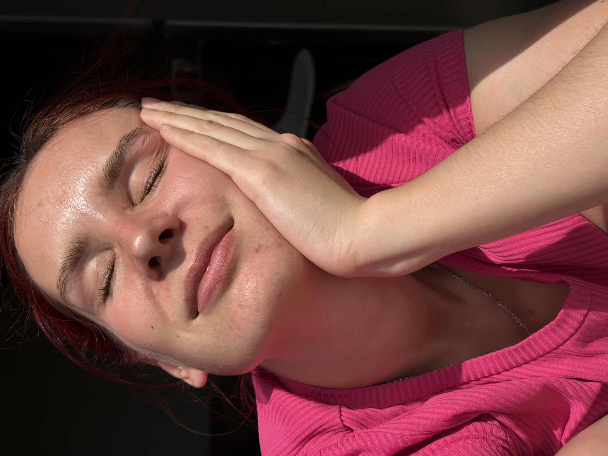 A woman is captured in an eye-level, close-up shot. Her eyes are closed, and one hand is resting on her cheek. She is wearing a pink ribbed top and a silver chain necklace. The background is dark, with a possible window or frame visible. The presence of red undertones in her hair can be detected, but the exact color can't be precisely stated without a colorimetric analysis. Based solely on the provided image, there is no information present that places the photo within the city of Chaikovsky, Russia.